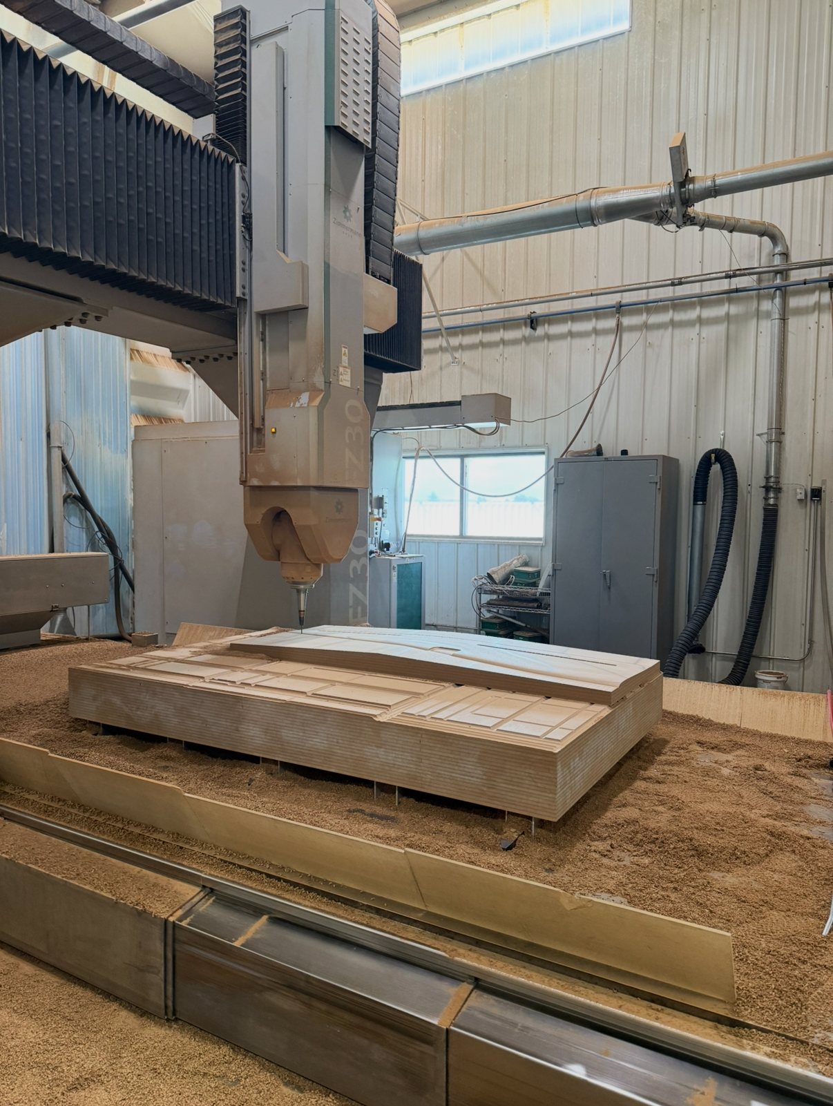 Industrial CNC machine working on wooden pieces in a workshop, with wood shavings and equipment around.