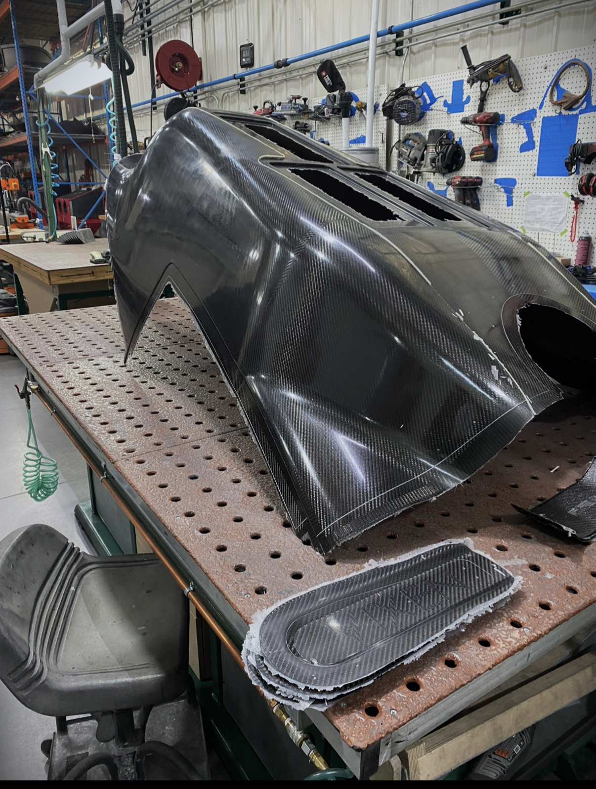 A carbon fiber car component, likely a hood or body panel, placed on a workbench with tools and equipment in the background of a workshop or garage