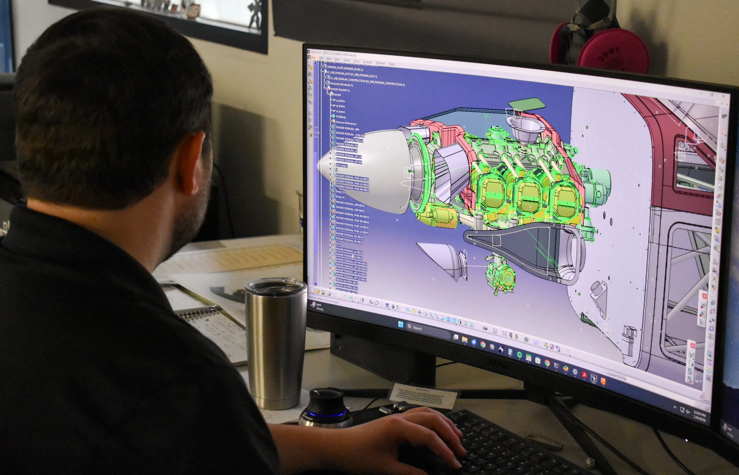 A person working on a CAD design of a vehicle engine on a computer monitor at a desk.