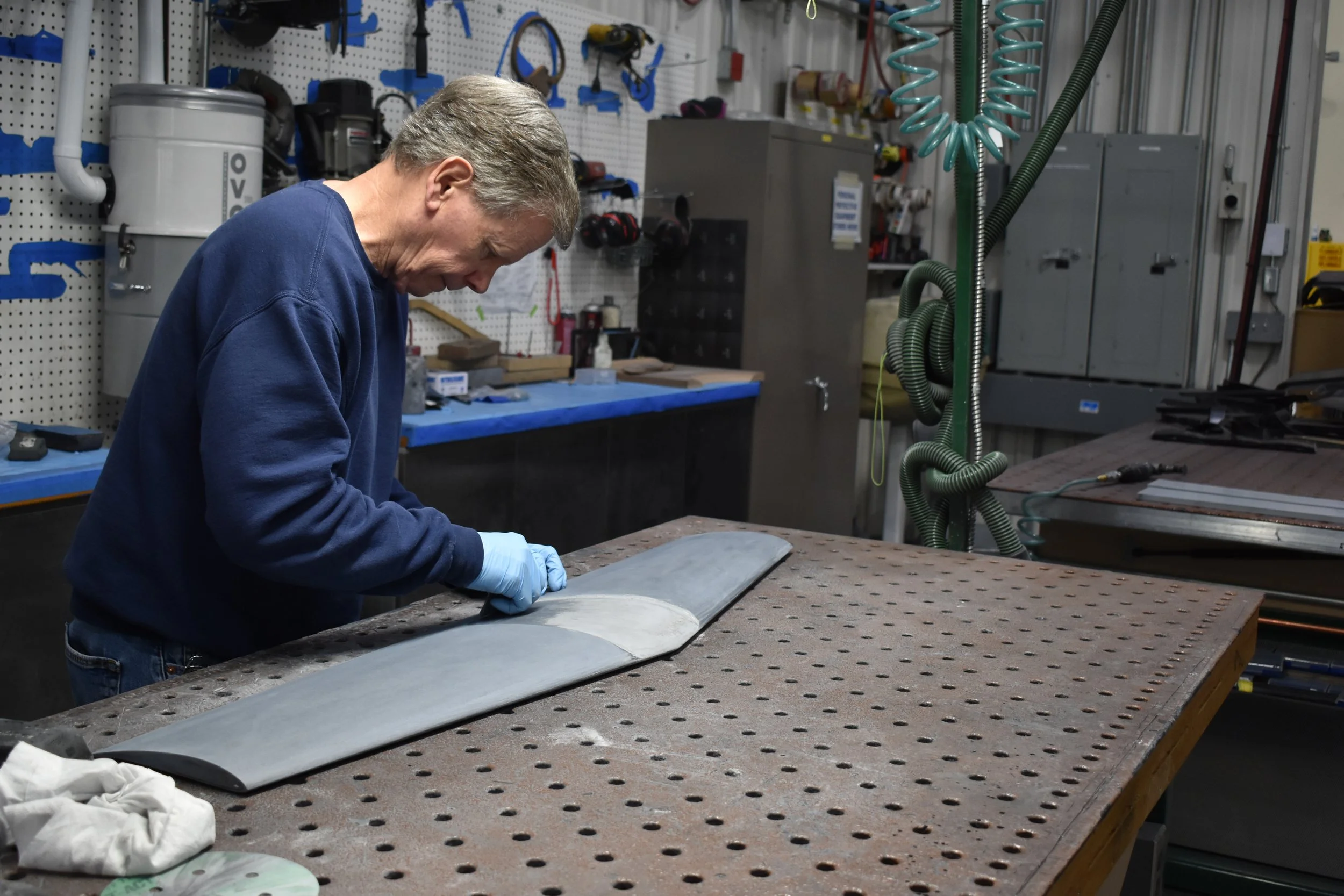 Man working on a large piece of metal in a workshop with tools hanging on pegboards and equipment in the background.