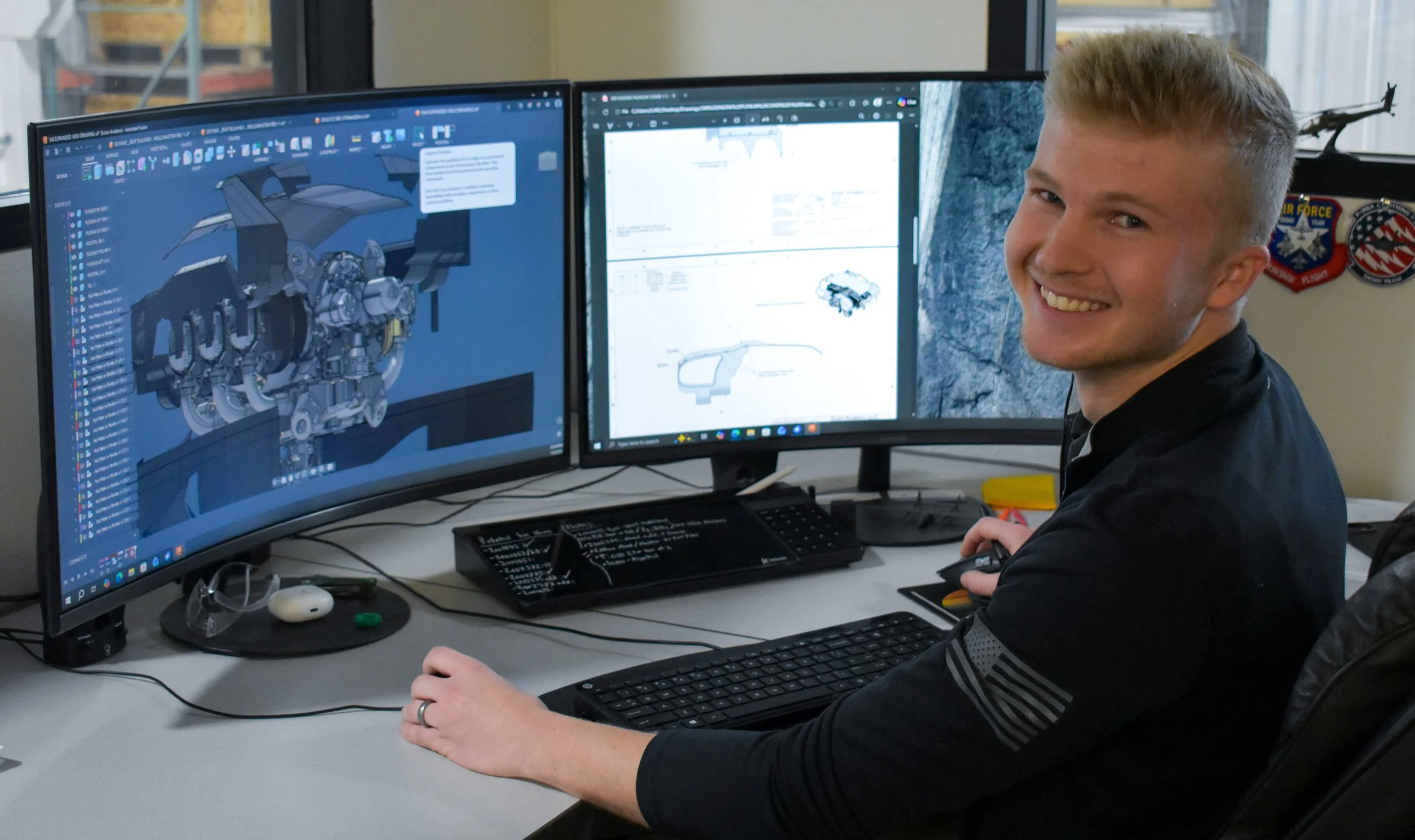 A young man sitting at a desk with three computer monitors, working on a 3D model of a car engine and a technical drawing. He is smiling and wearing a black shirt, with a keyboard and mouse in front of him.