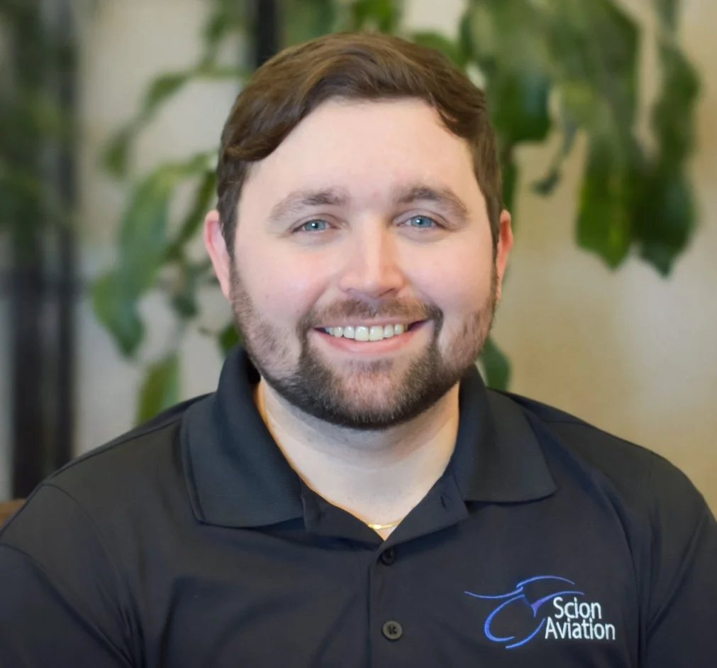 A smiling man with short brown hair and a beard, wearing a black polo shirt with a blue logo that says 'Scion Aviation', sitting in front of blurred green plants.