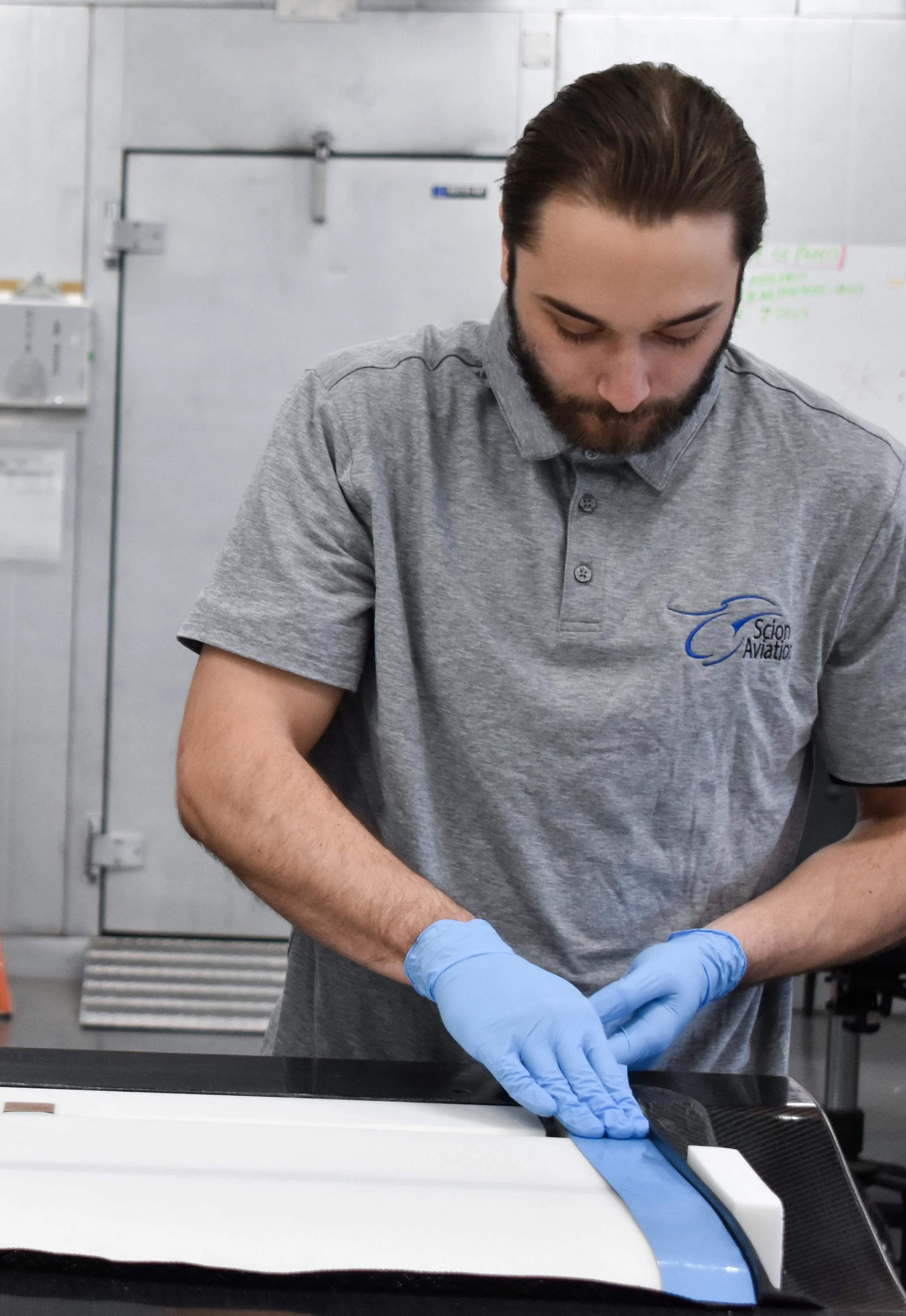 Man wearing a gray polo shirt with a logo and blue gloves working on a black motorized aircraft component in a workshop.