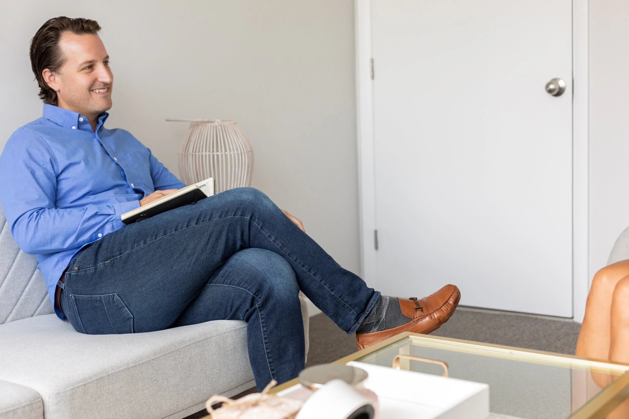 A man sitting on a light gray sofa holding a notebook, smiling, in a room with white walls and door, with a lamp and coffee table nearby.