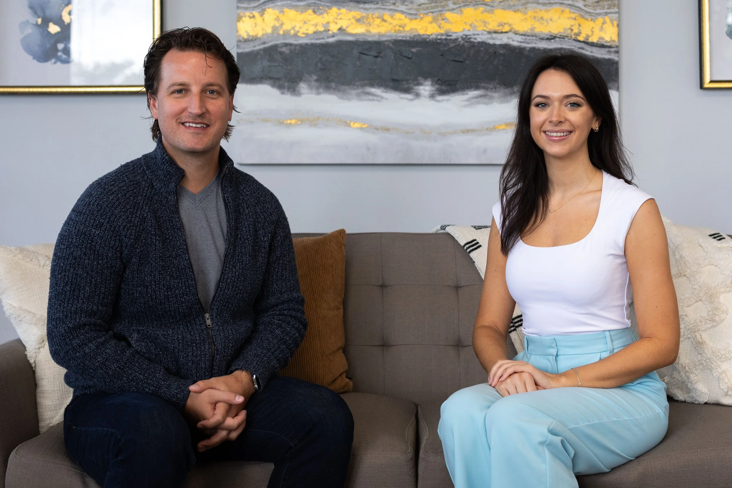 A man and woman are sitting on a sofa in the lobby of the Florida Relationship Institute, smiling at the camera. The man is wearing a dark blue sweater over a gray shirt, and the woman is dressed in a white top and light blue pants.