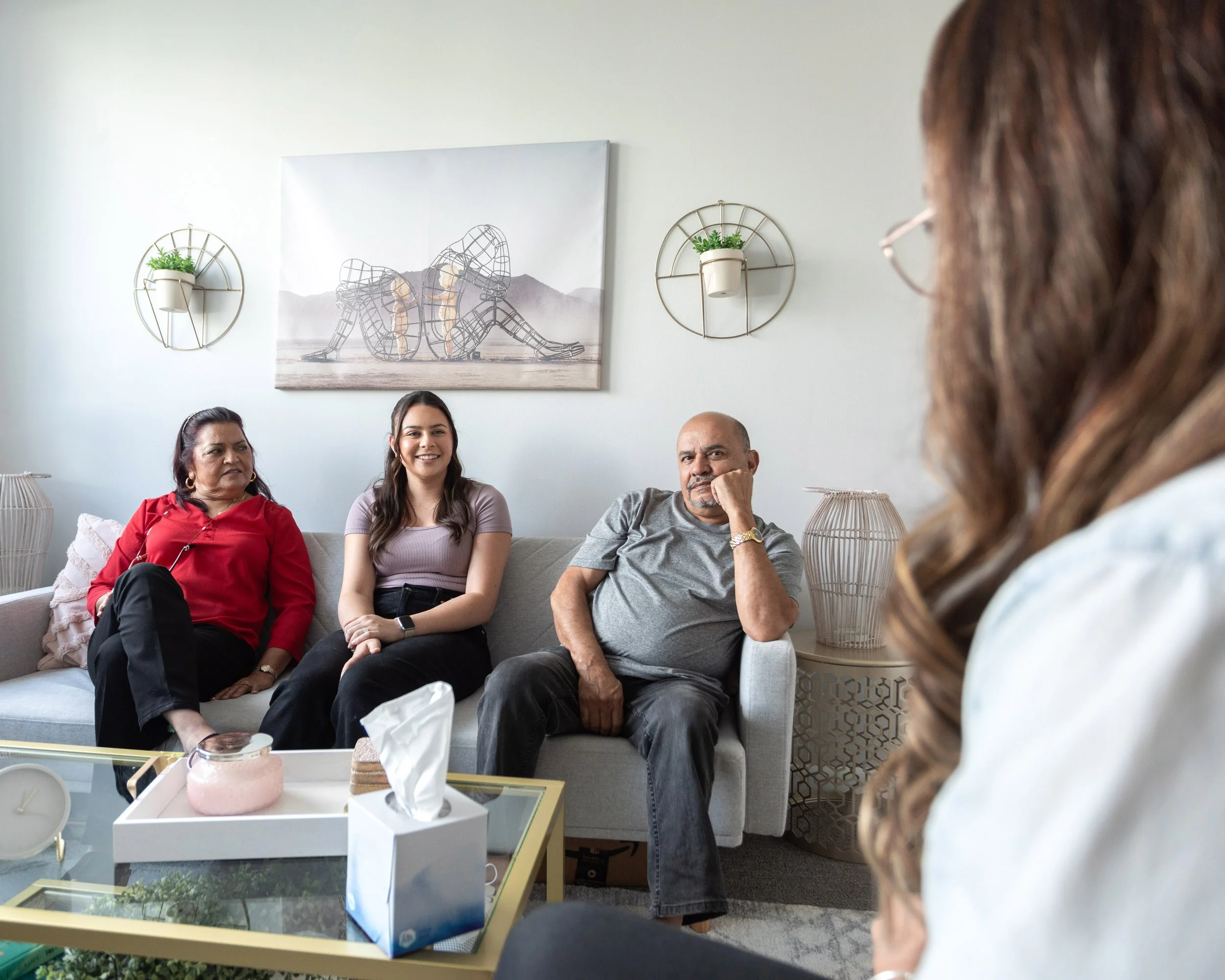 A group of four people at the Florida Relationship Institute are engaging in a conversation. Three are seated on a light-colored sofa, including a woman in red, a woman in lavender, and a man in gray. A therapist is seated and listening.
