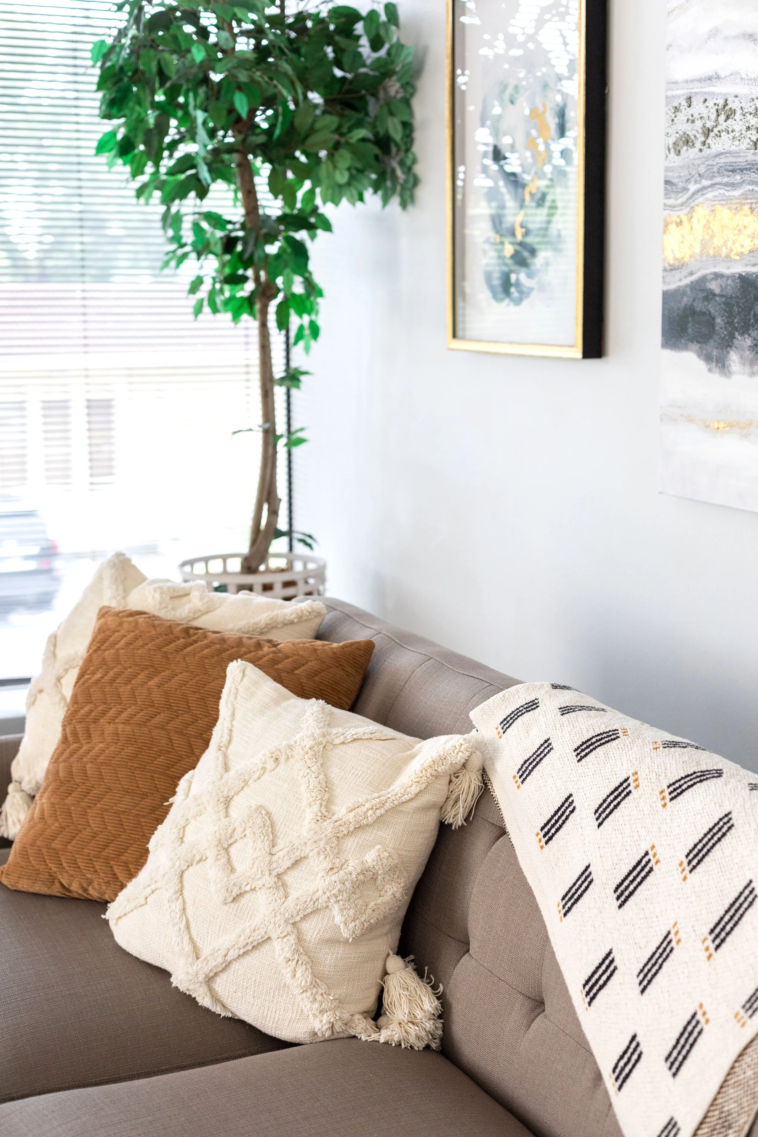 Therapy room corner with a beige sofa adorned with decorative pillows, including a brown, a cream with tassels, and a white pillow with black geometric patterns. A large green potted plant, a window with blinds, and framed artwork hang on the wall.