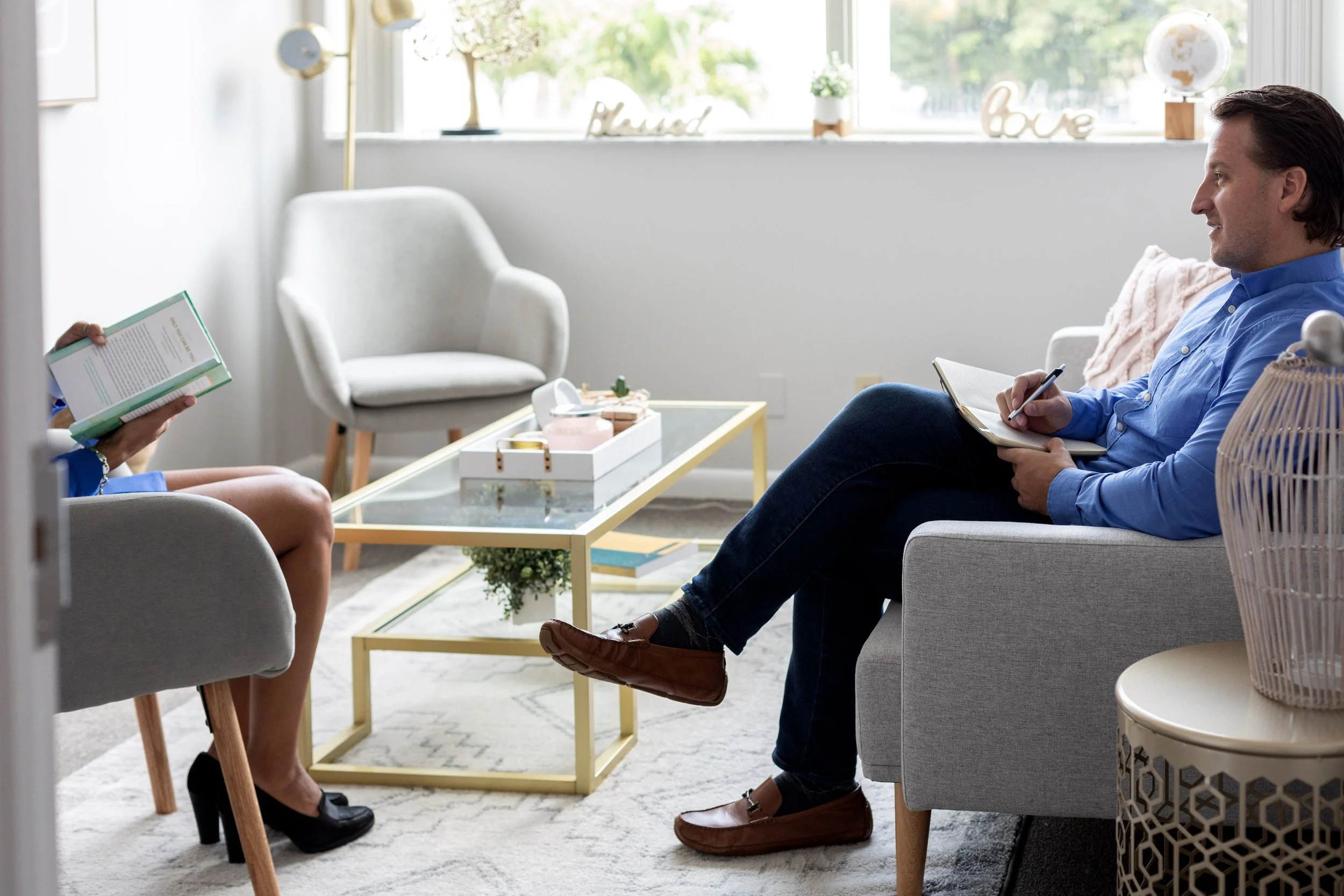 A therapist and patient having a session in a bright, modern office with a large window, decorative words on the windowsill, and a coffee table with plants and books.