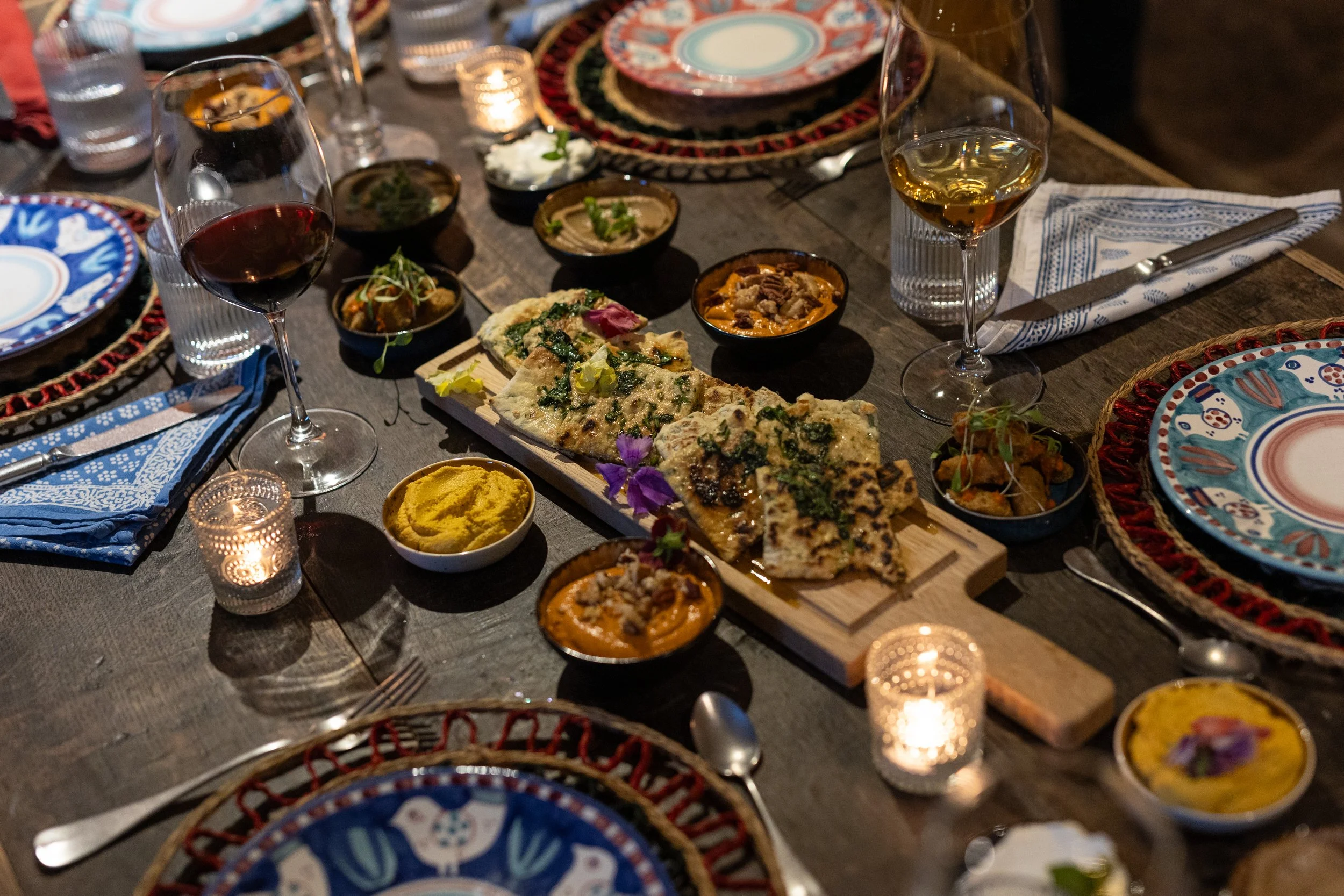 A dinner table set with colorful plates, glasses of red and white wine, various small bowls of Indian dishes, a wooden serving board with naan bread, and lit candles.