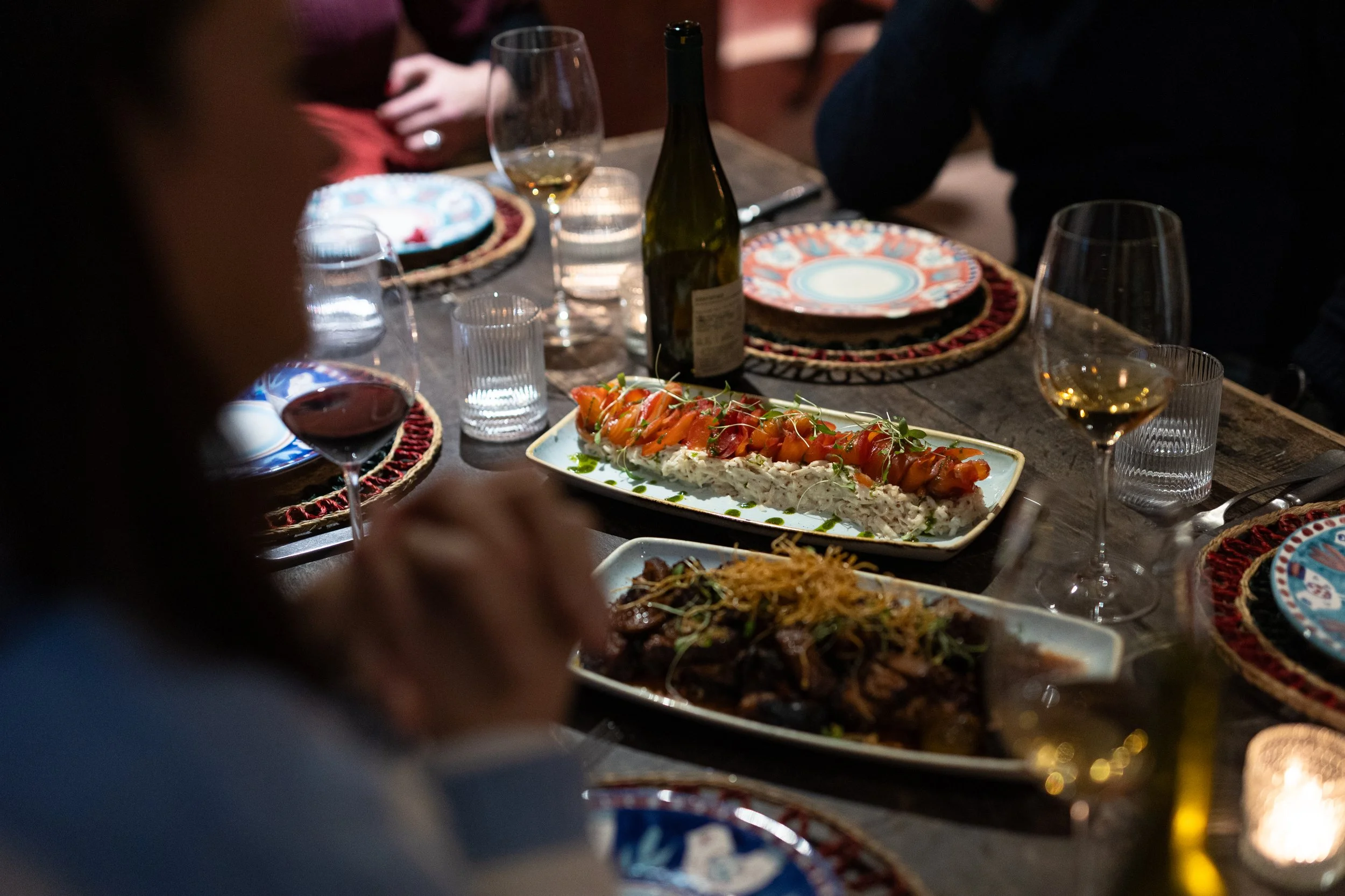 A dinner table with plates, glasses of white wine, water, a bottle of wine, and dishes including a salmon appetizer topped with vegetables and a cooked meat dish. The setting appears to be a cozy, dimly-lit dining environment with festive tableware.
