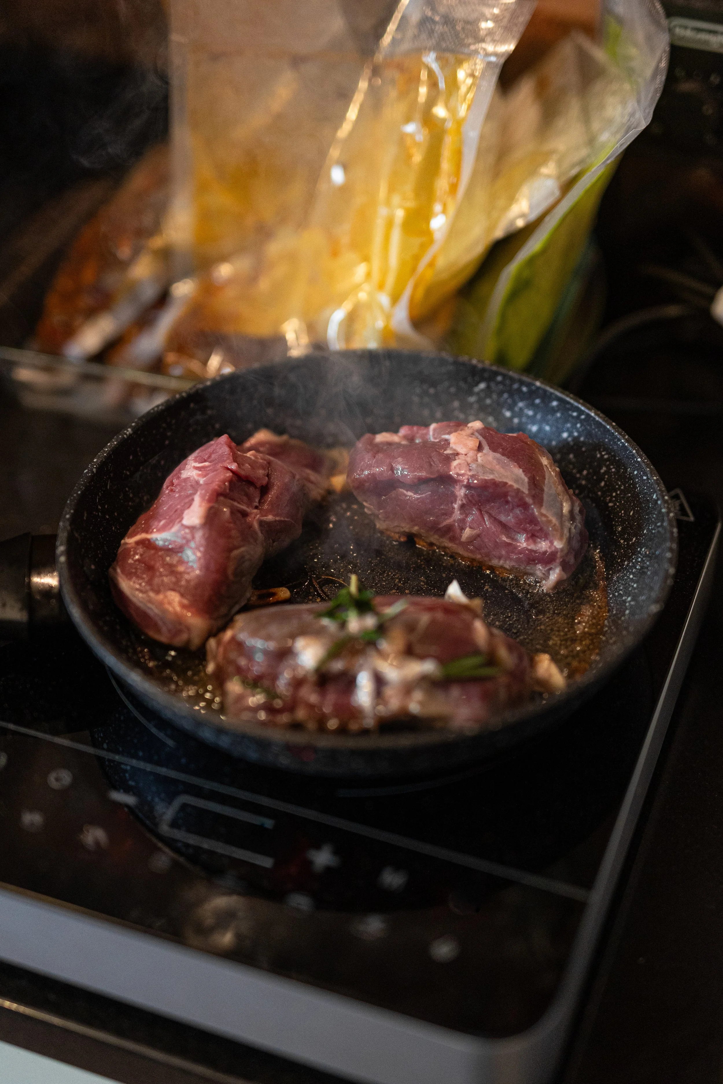 Three raw pieces of beef cooking in a frying pan with some herbs and garlic, on a stove. In the background, there is a bag of yellow items, possibly potatoes or onions.