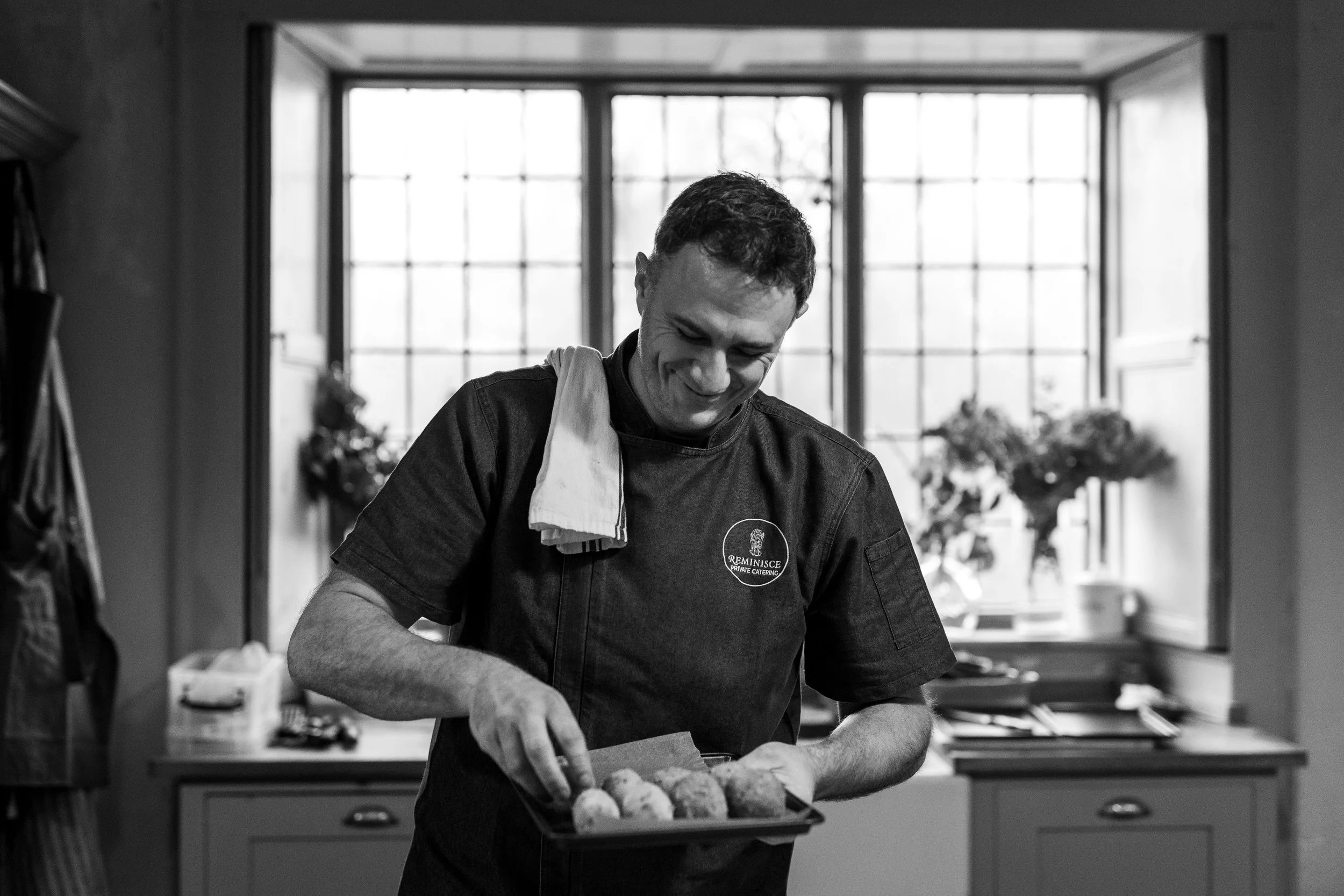 A man in a chef's uniform holding a tray of food, smiling, in a kitchen with large windows in the background.