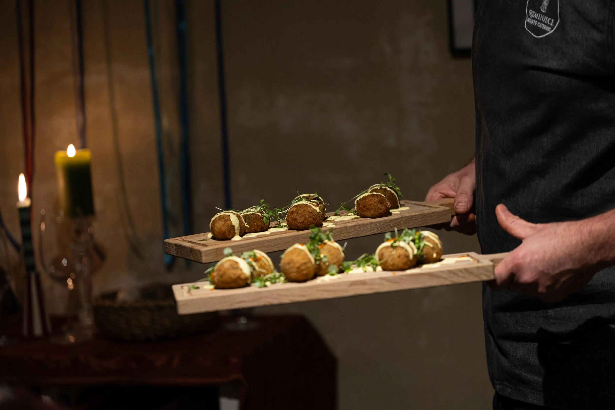 Person holding two wooden serving boards with fried food garnished with greens, in a dimly lit setting with candles in the background.