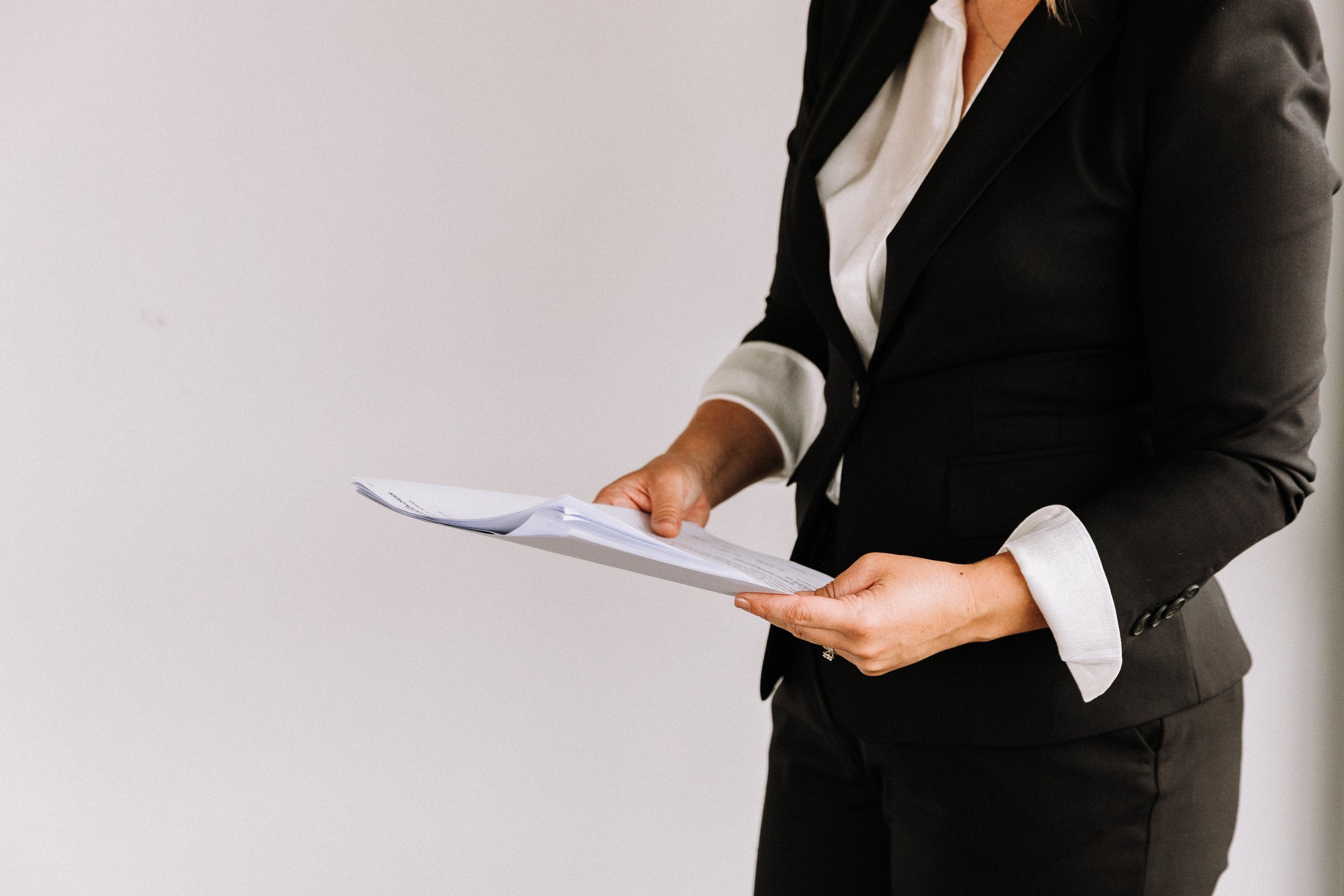 A woman dressed in a black suit with a white shirt, holding a stack of papers.