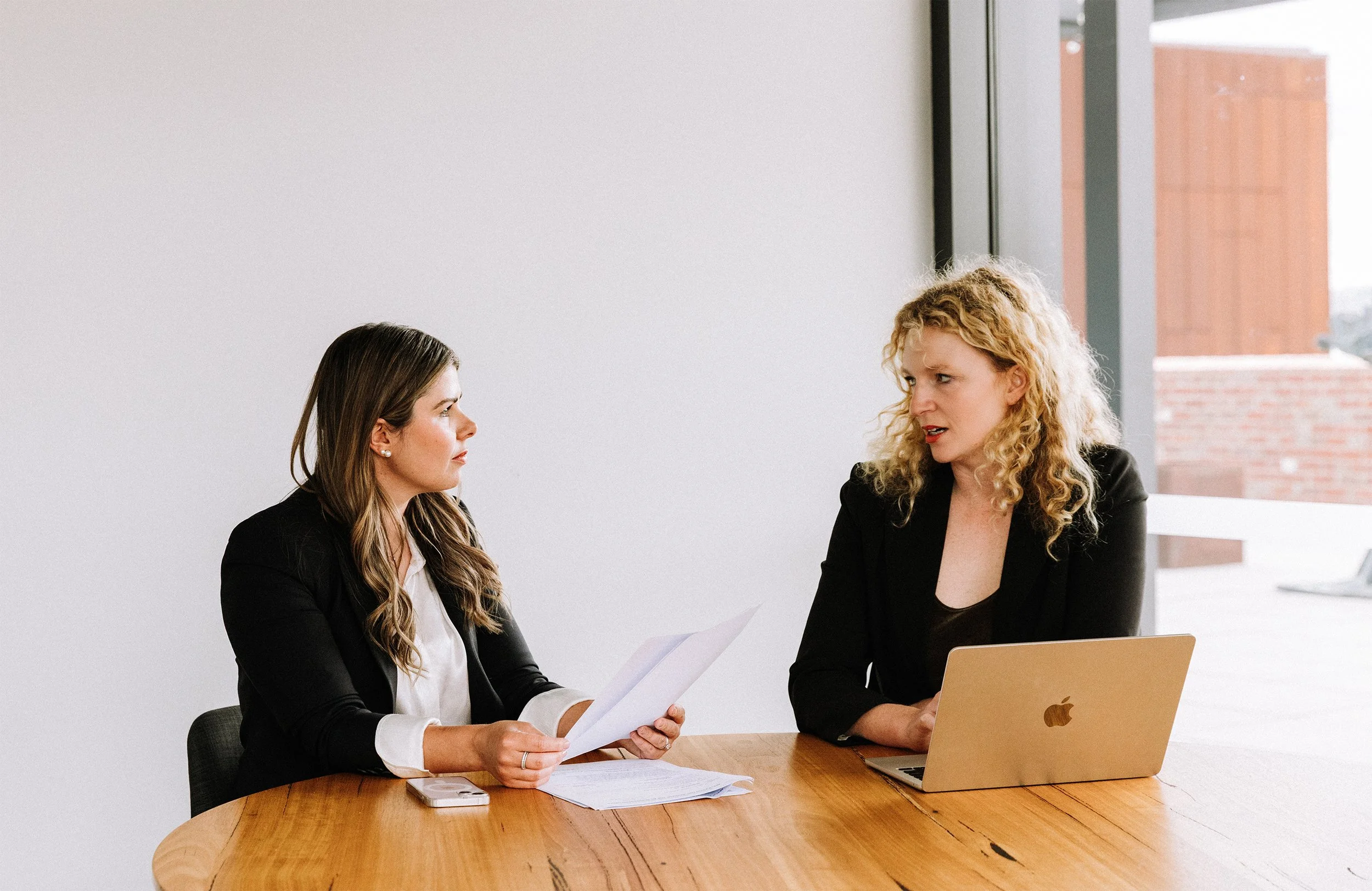 Two women sitting at a round wooden table in a modern office, engaging in a conversation. One woman has long brown hair, wearing a black blazer, holding papers. The other woman has curly blonde hair, wearing a black blazer, using a silver MacBook laptop.