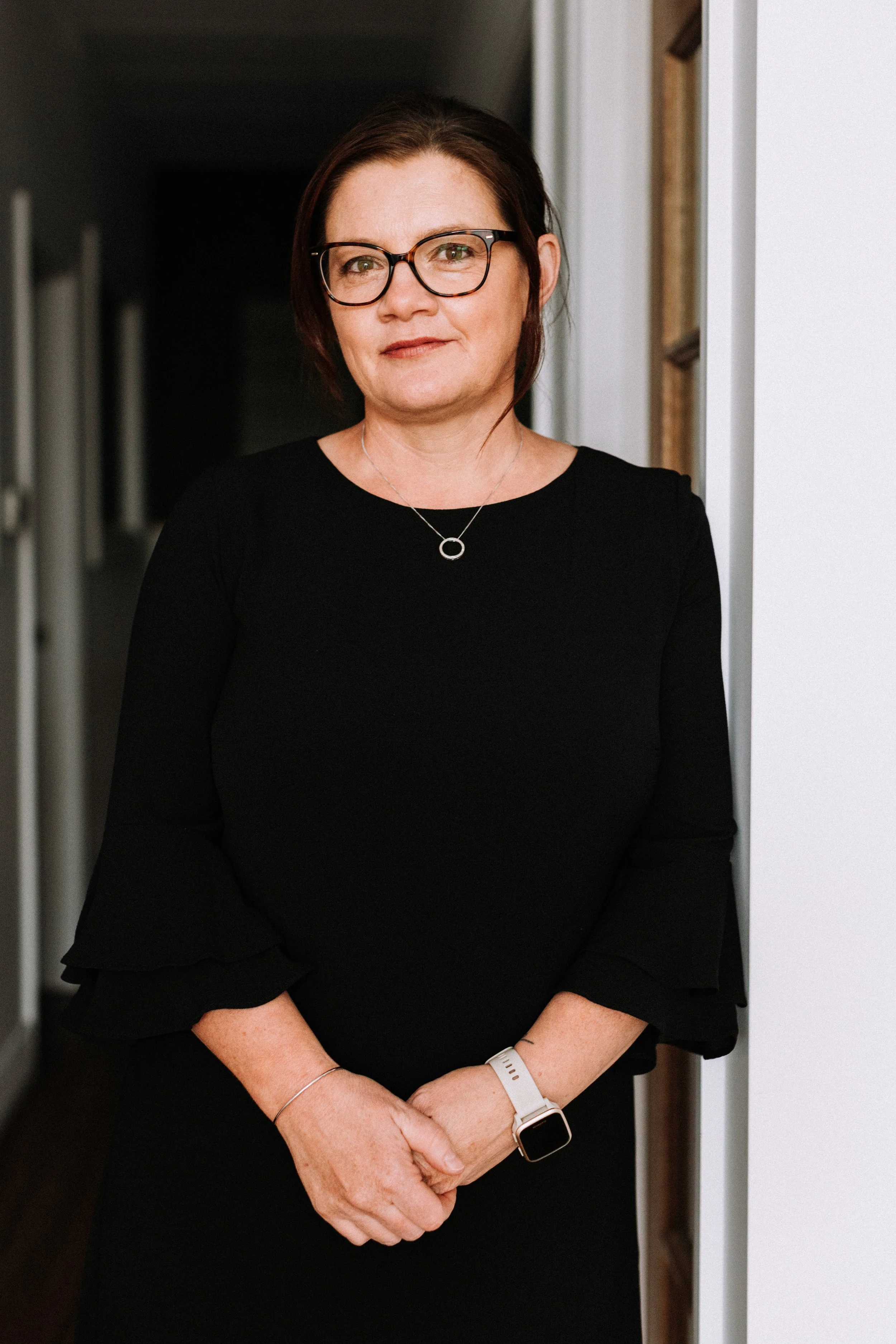 A woman with short dark hair, glasses, and a black dress standing in a hallway.