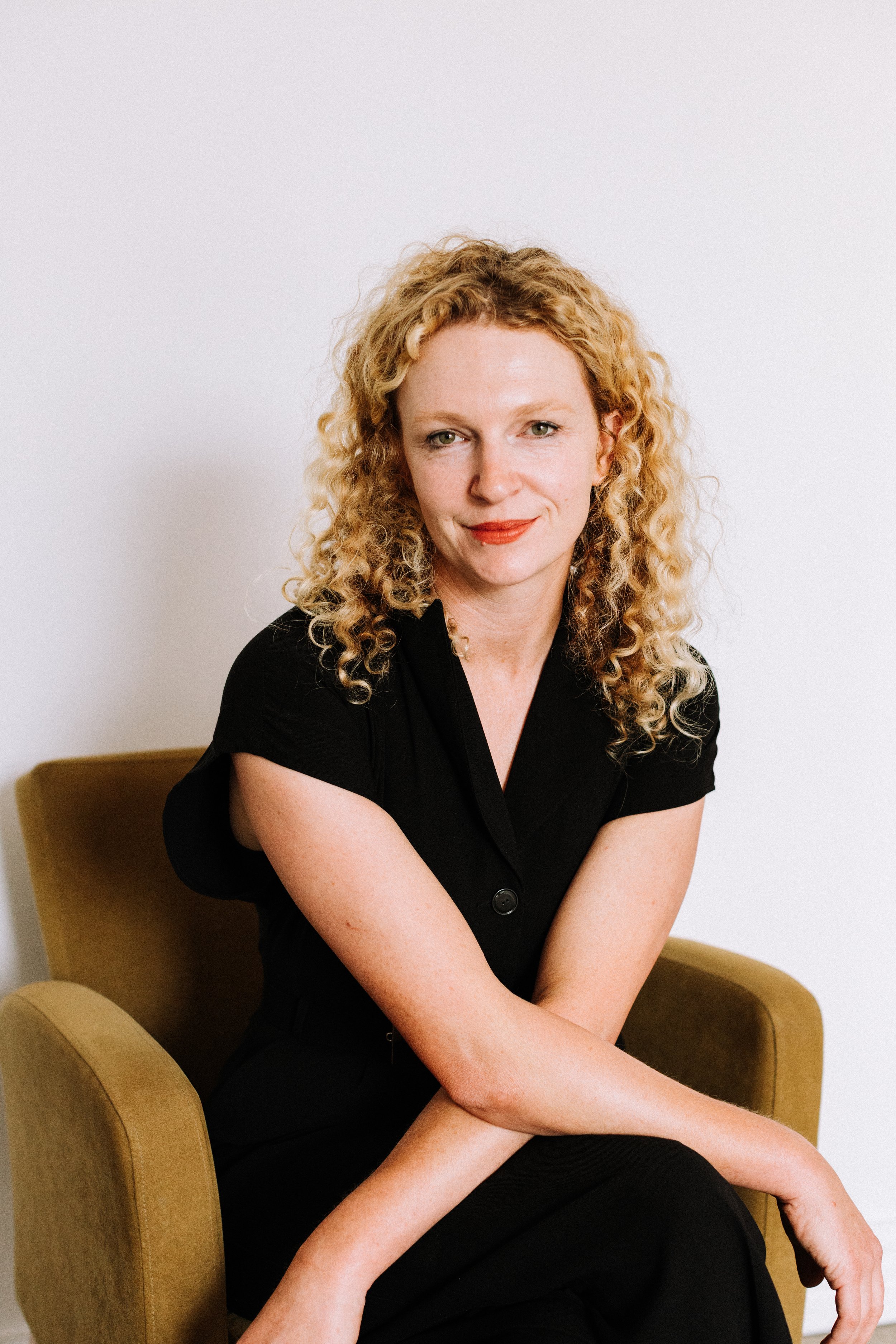 A woman with curly blonde hair sitting on a green armchair against a plain white wall, wearing a black dress and red lipstick.