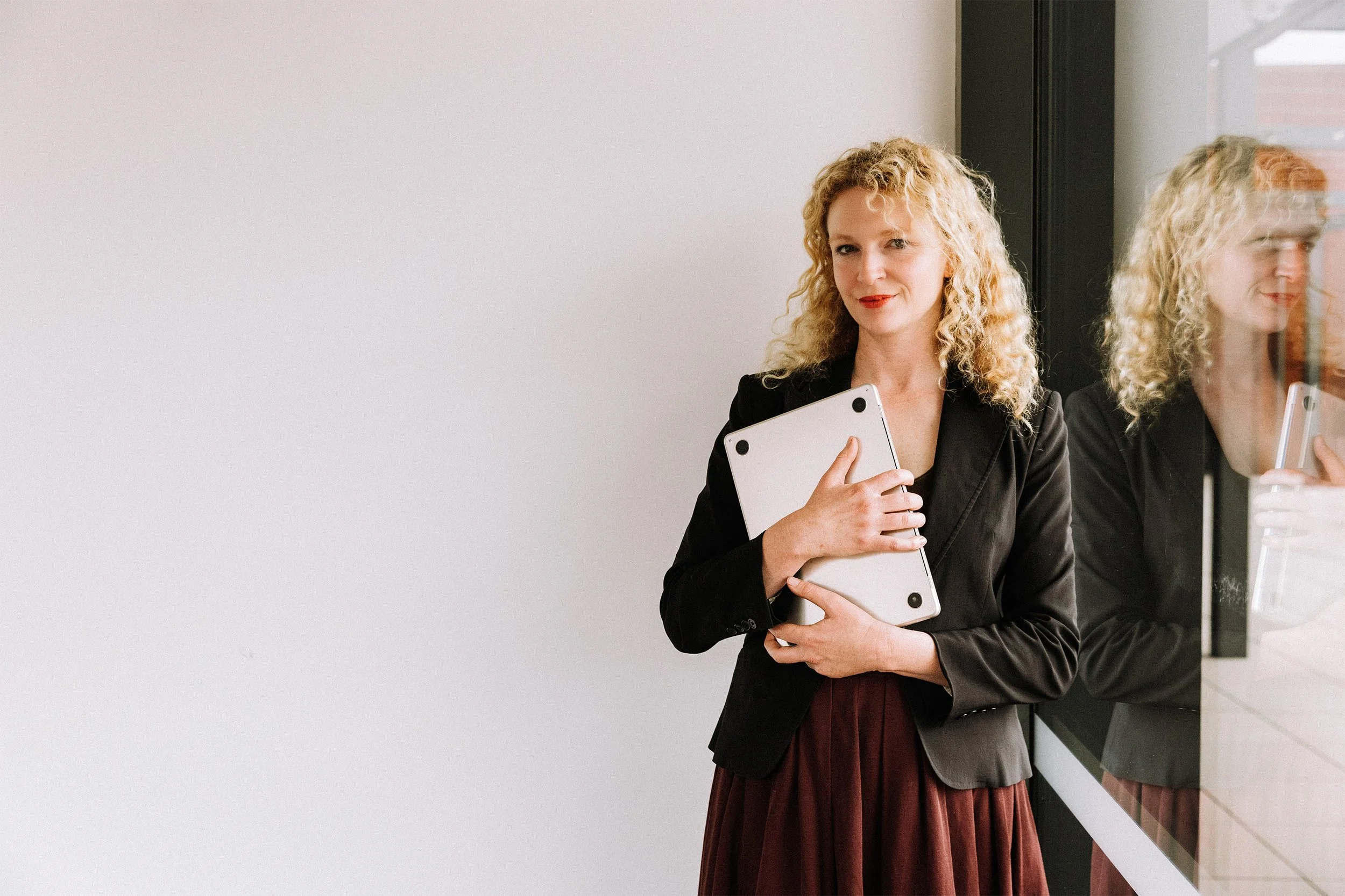 A woman with curly blonde hair in a black blazer, holding a silver laptop, stands next to a window with her reflection visible in the glass.