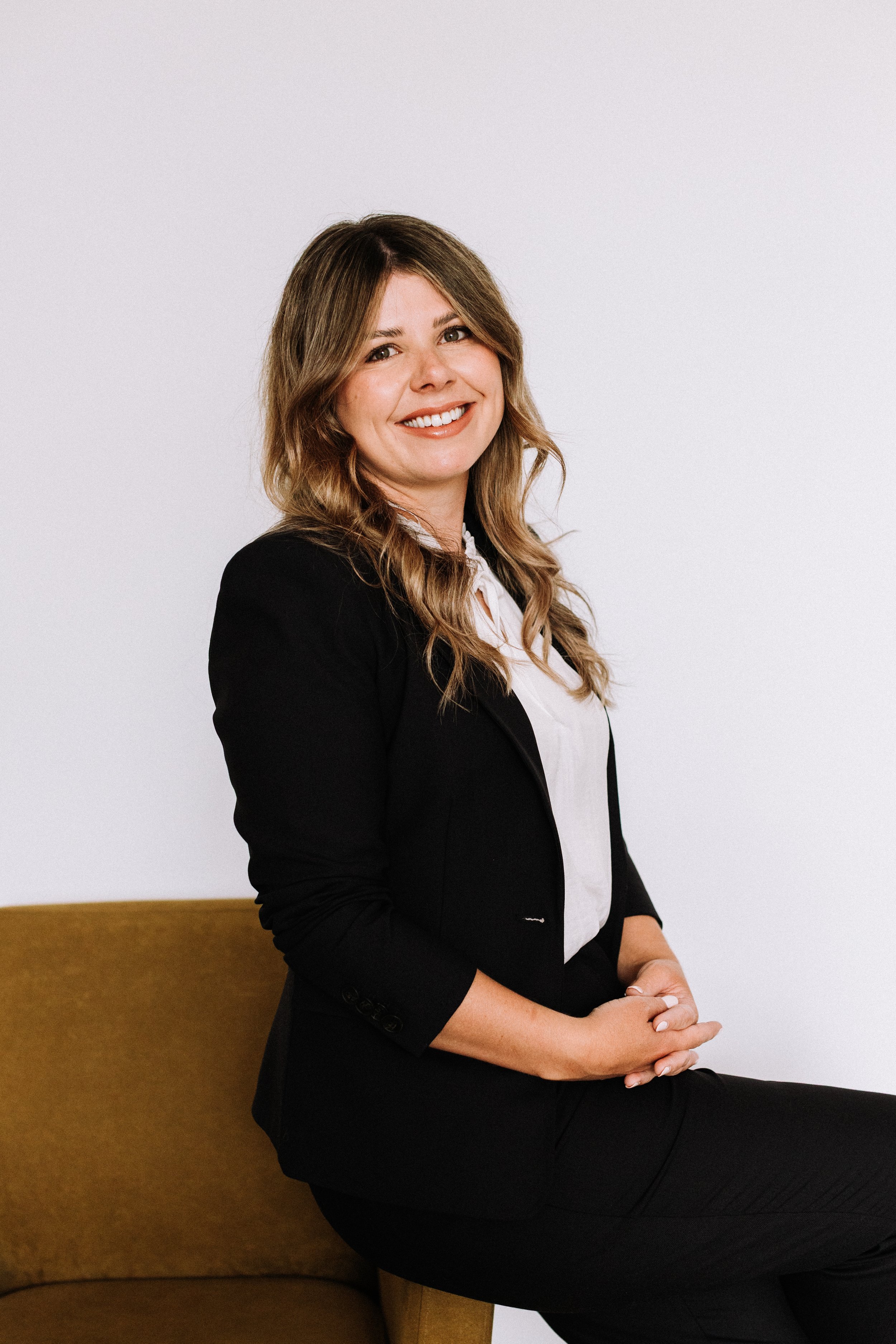 A woman with wavy brown hair smiling, sitting on a brown couch, wearing a black blazer and white blouse against a plain white background.