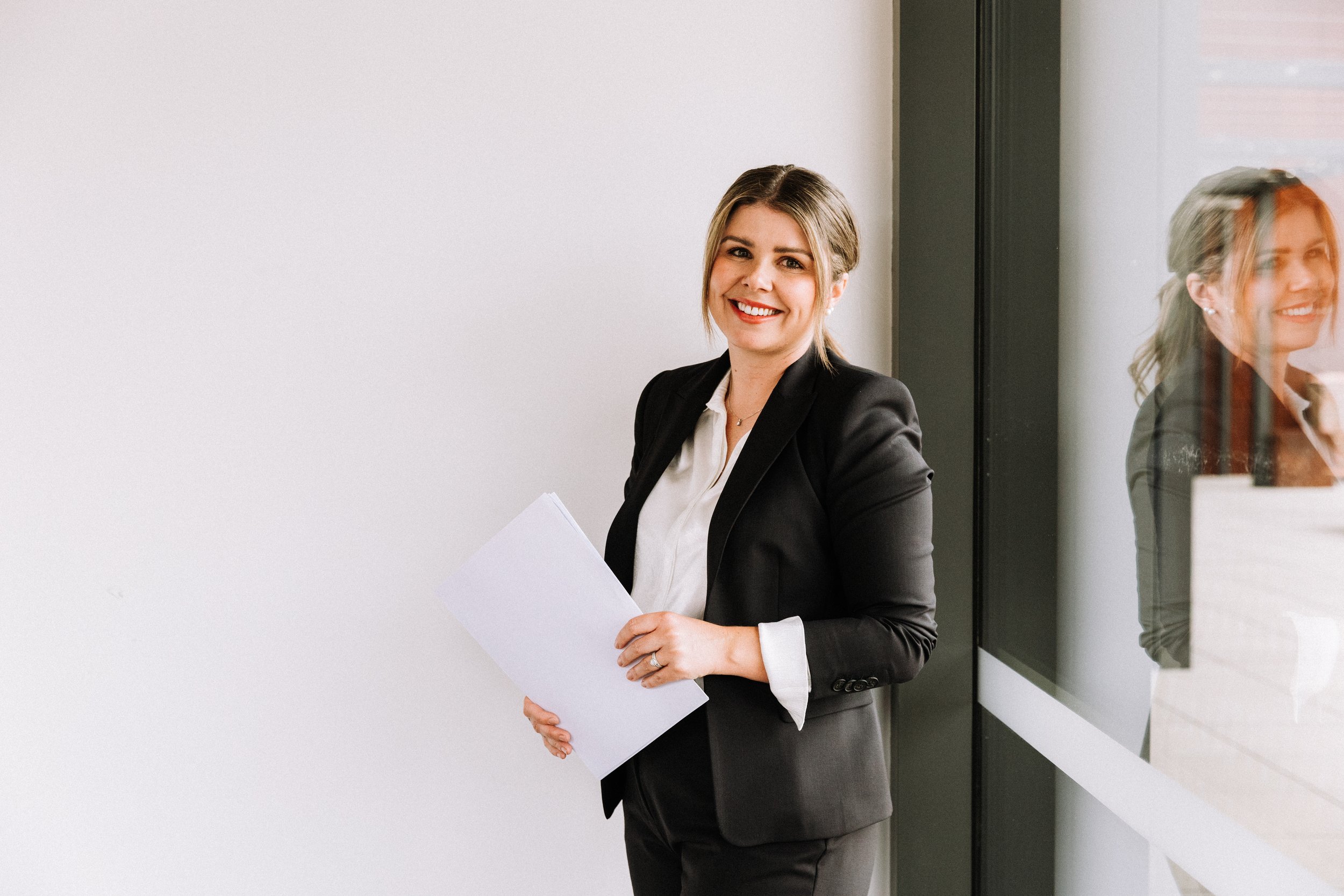 A smiling woman in a black blazer and white shirt holding papers, standing by a glass wall with her reflection visible.