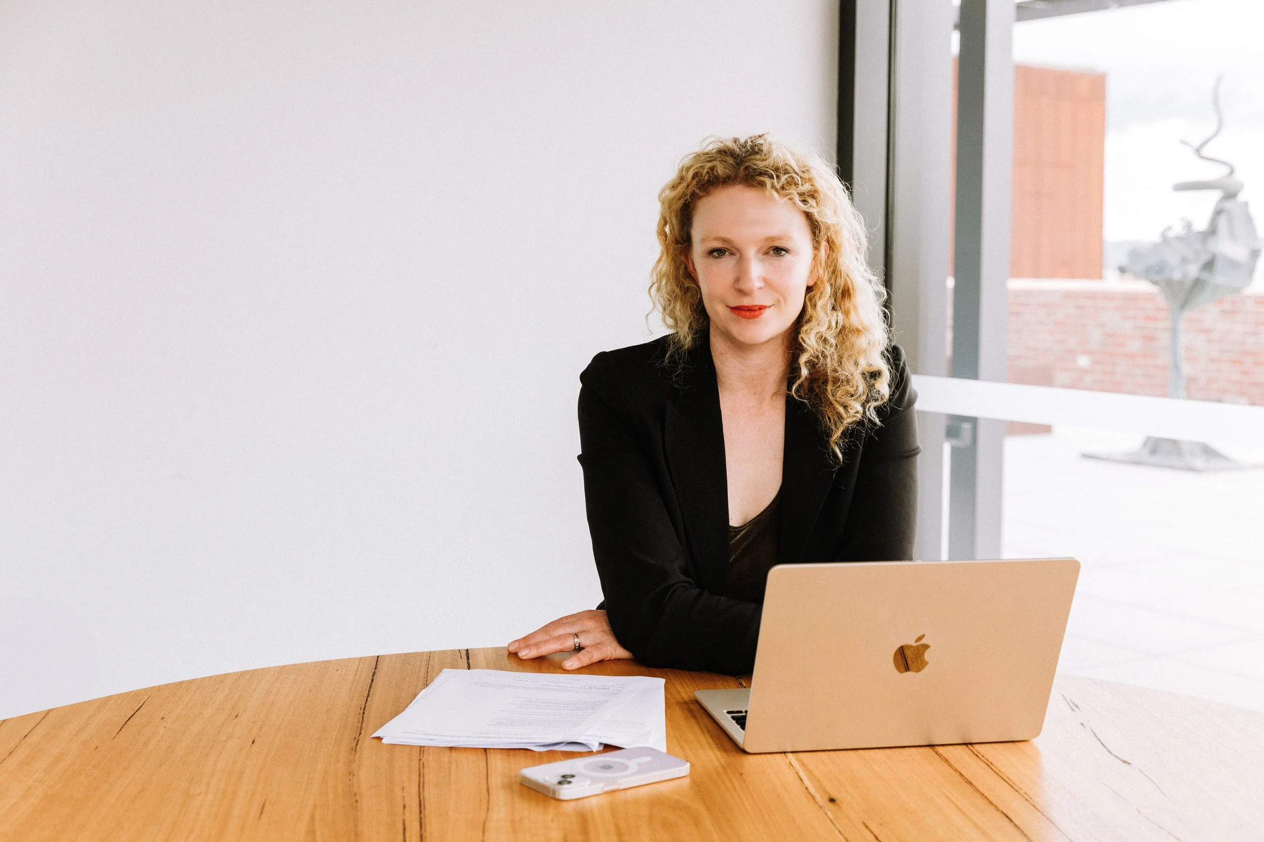 A woman with curly blond hair sitting at a wooden table with a laptop, papers, and a smartphone, in a modern office with large windows.