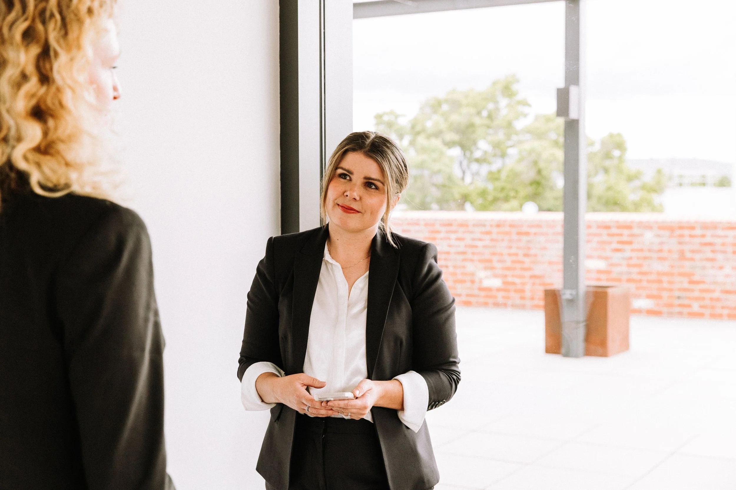Two women in business attire engaging in conversation in a modern office space with large windows, outdoor view, and brick wall.