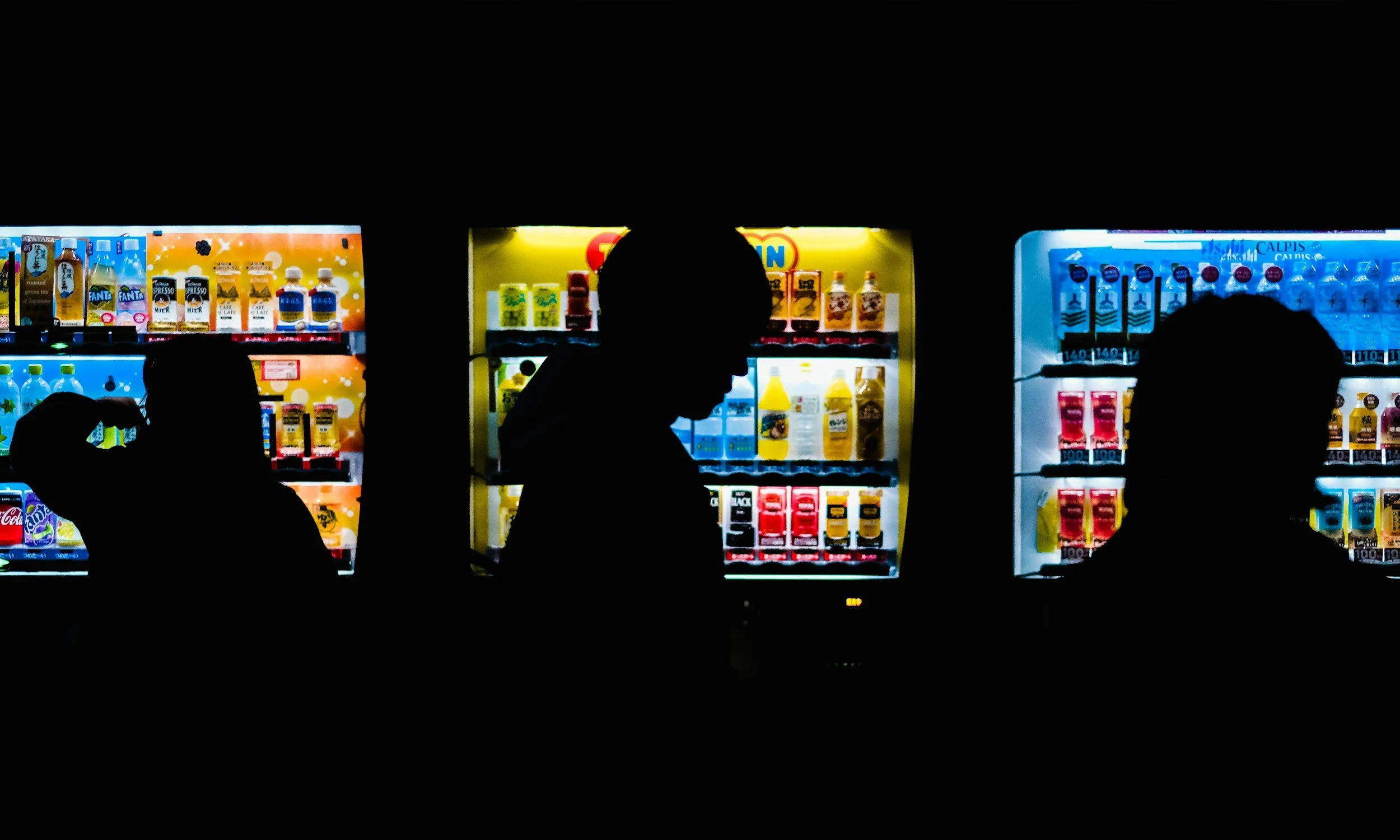Silhouettes of three people in front of illuminated vending machines with drinks and snacks.