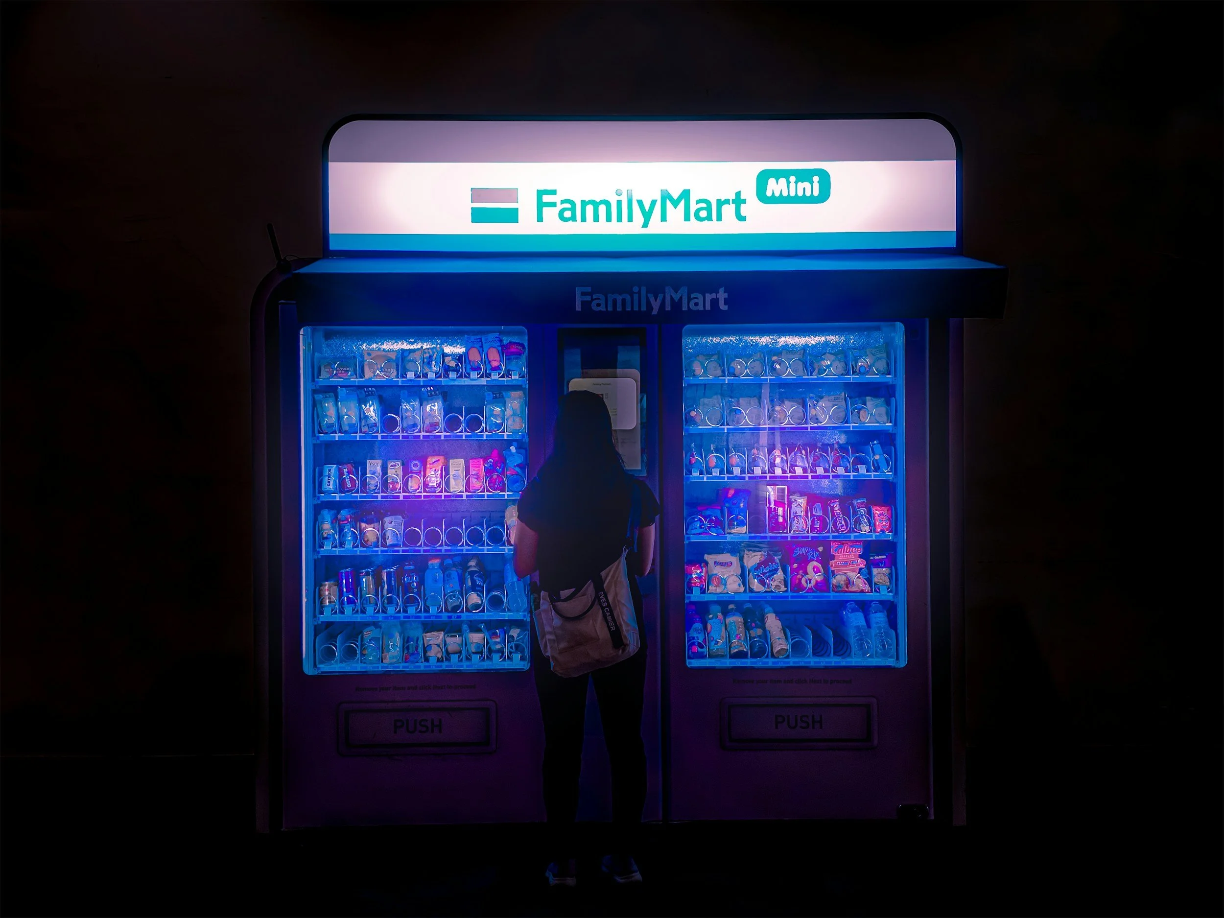 Person standing in front of a FamilyMart Mini vending machine illuminated with blue lighting, stocked with snacks and beverages.