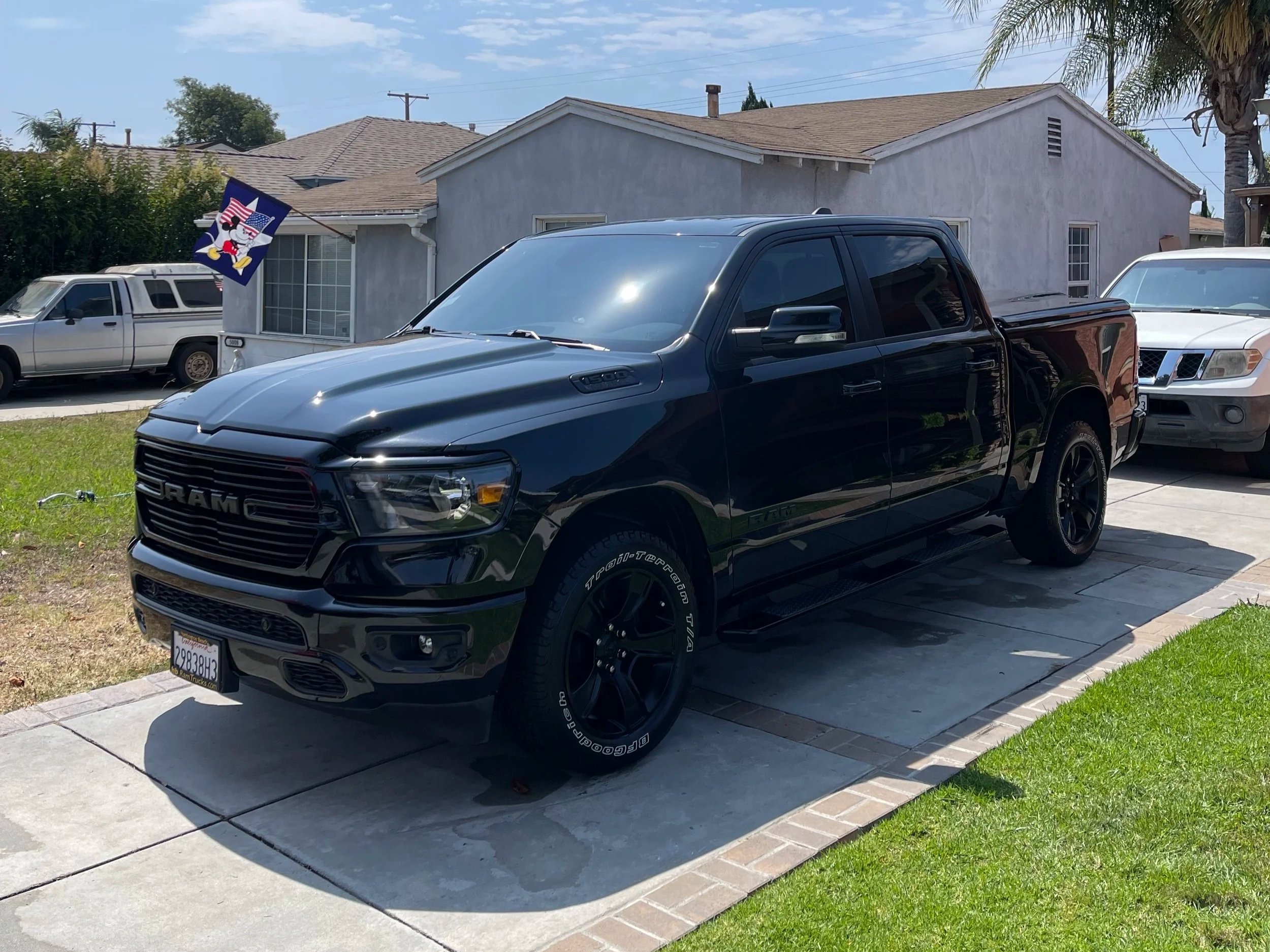 Black RAM pickup truck parked in driveway next to a green lawn, with a house, another truck, and a flag featuring a cartoon character in the background.