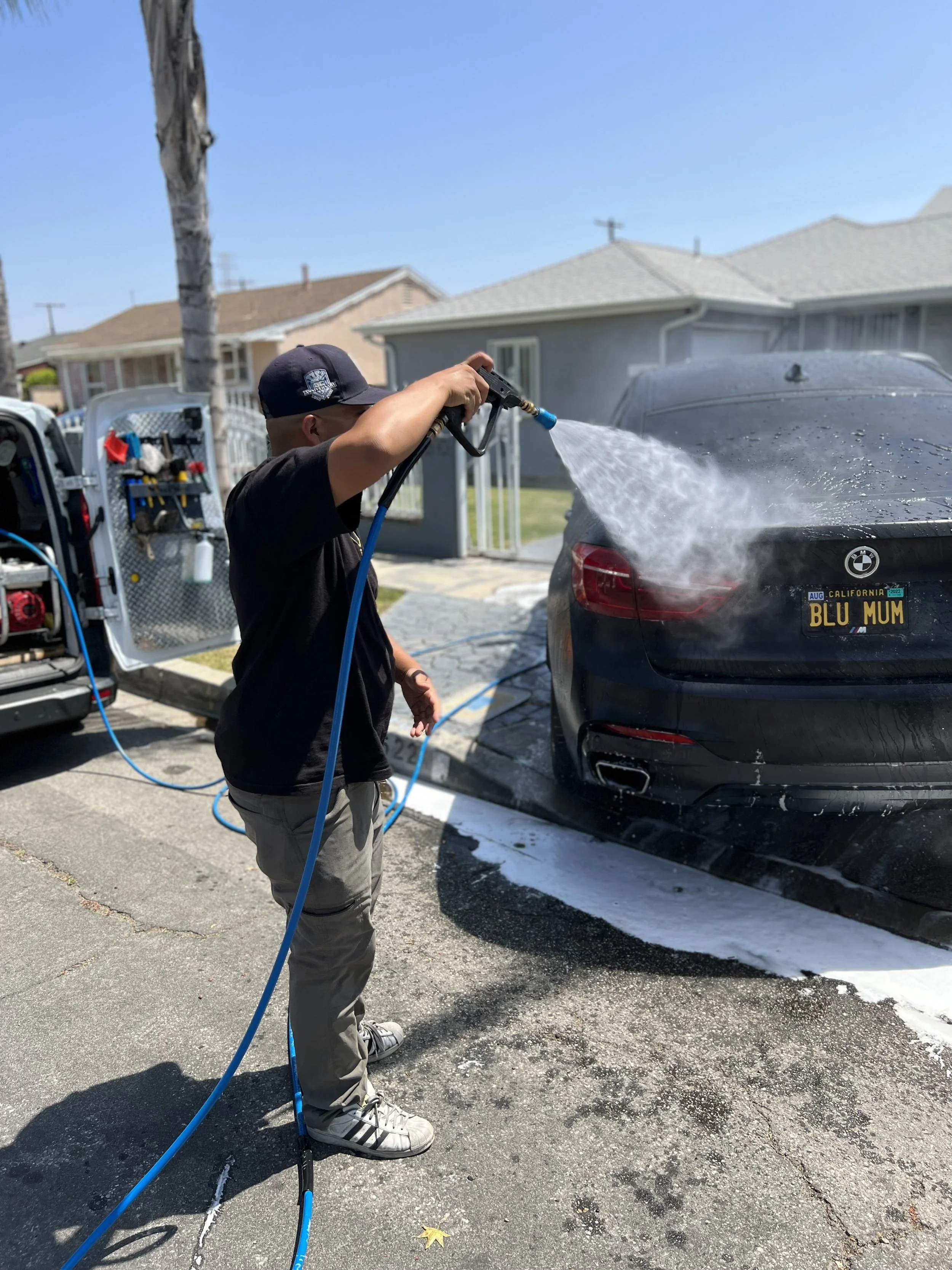 Mobile auto detailer rinsing a black BMW with a pressure washer during a foam wash on a sunny day