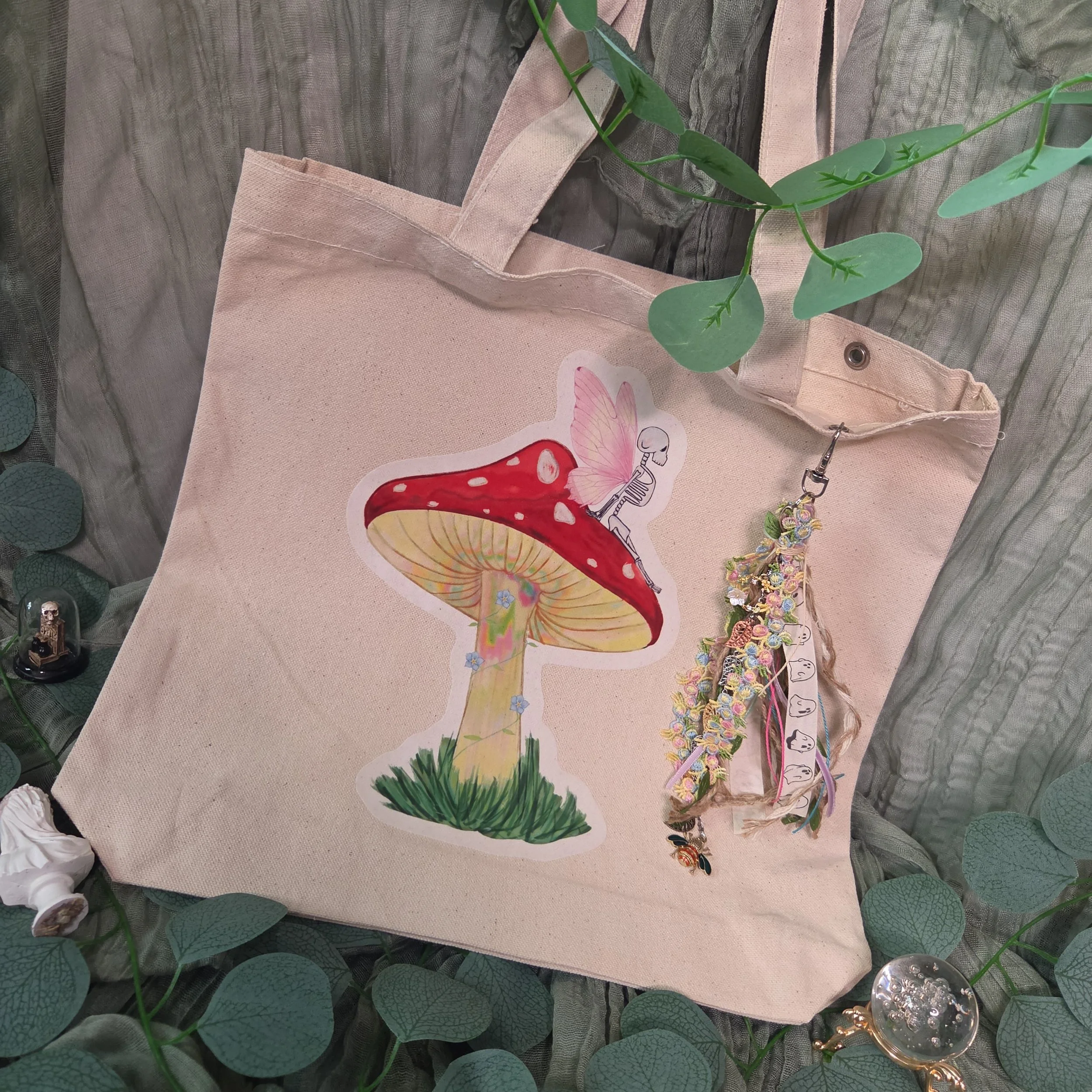 Drawing of a skeleton fairy sitting on a mushroom printed on a canvas tote bag with charm attached