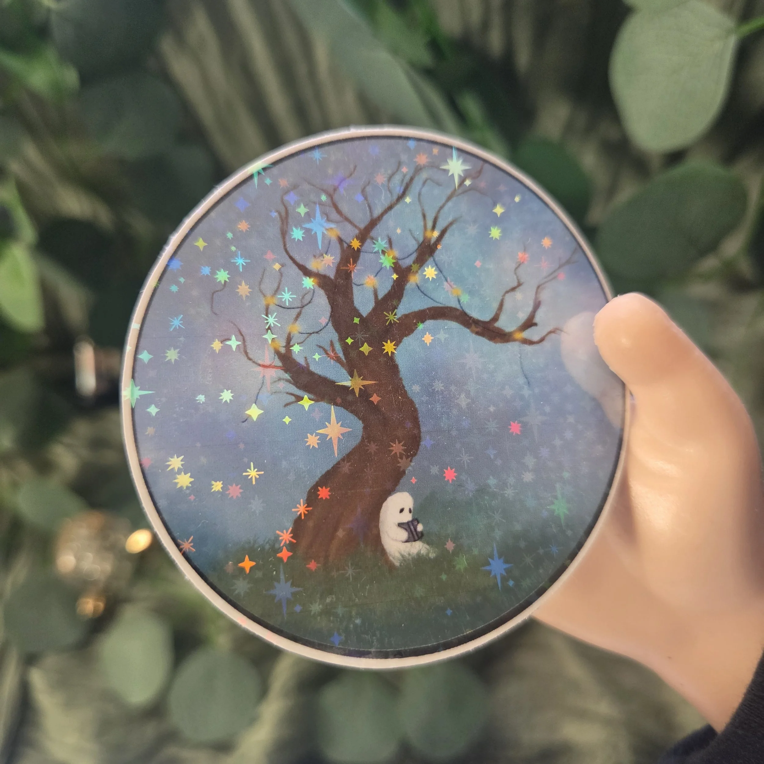 Hand holding a sticker of a small ghost reading beneath a tree at night with glowing lights in the branches and a purple-blue night sky (Copy)