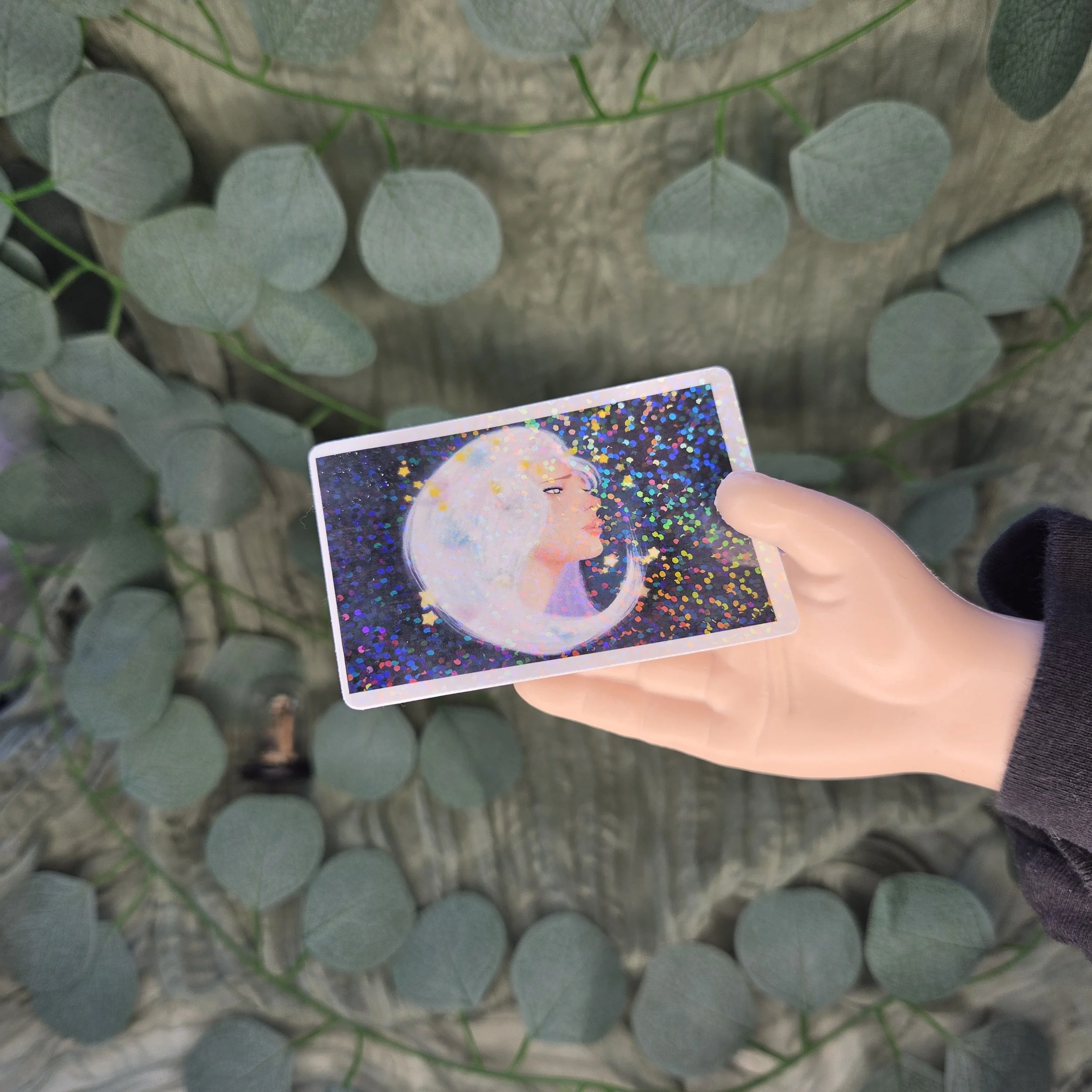A hand holding a drawing of a woman with flowing hair shaped like the moon, surrounded by glowing stars against a dark night sky with a holographic laminate.