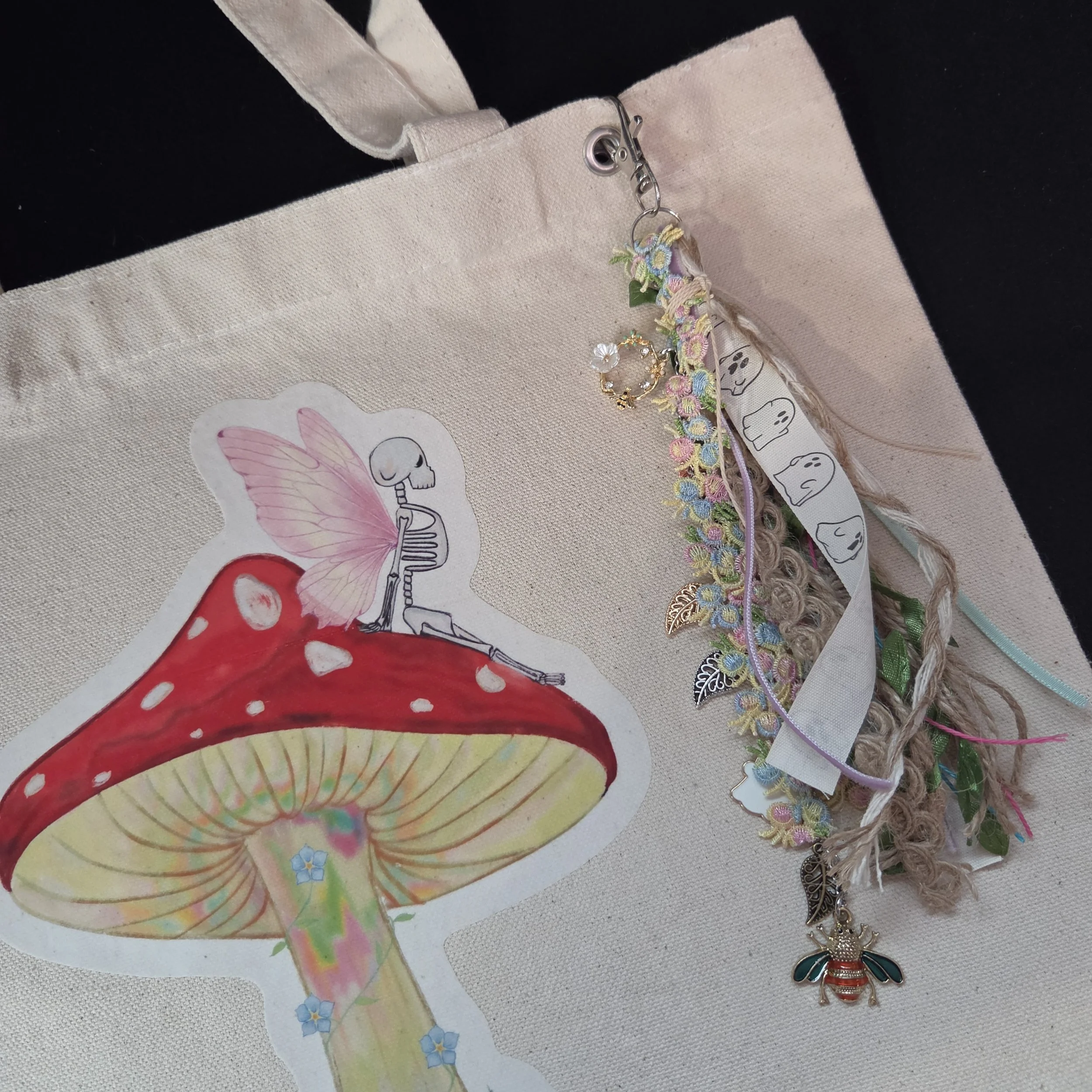 Drawing of a skeleton fairy sitting on a mushroom printed on a canvas tote bag with charm attached
