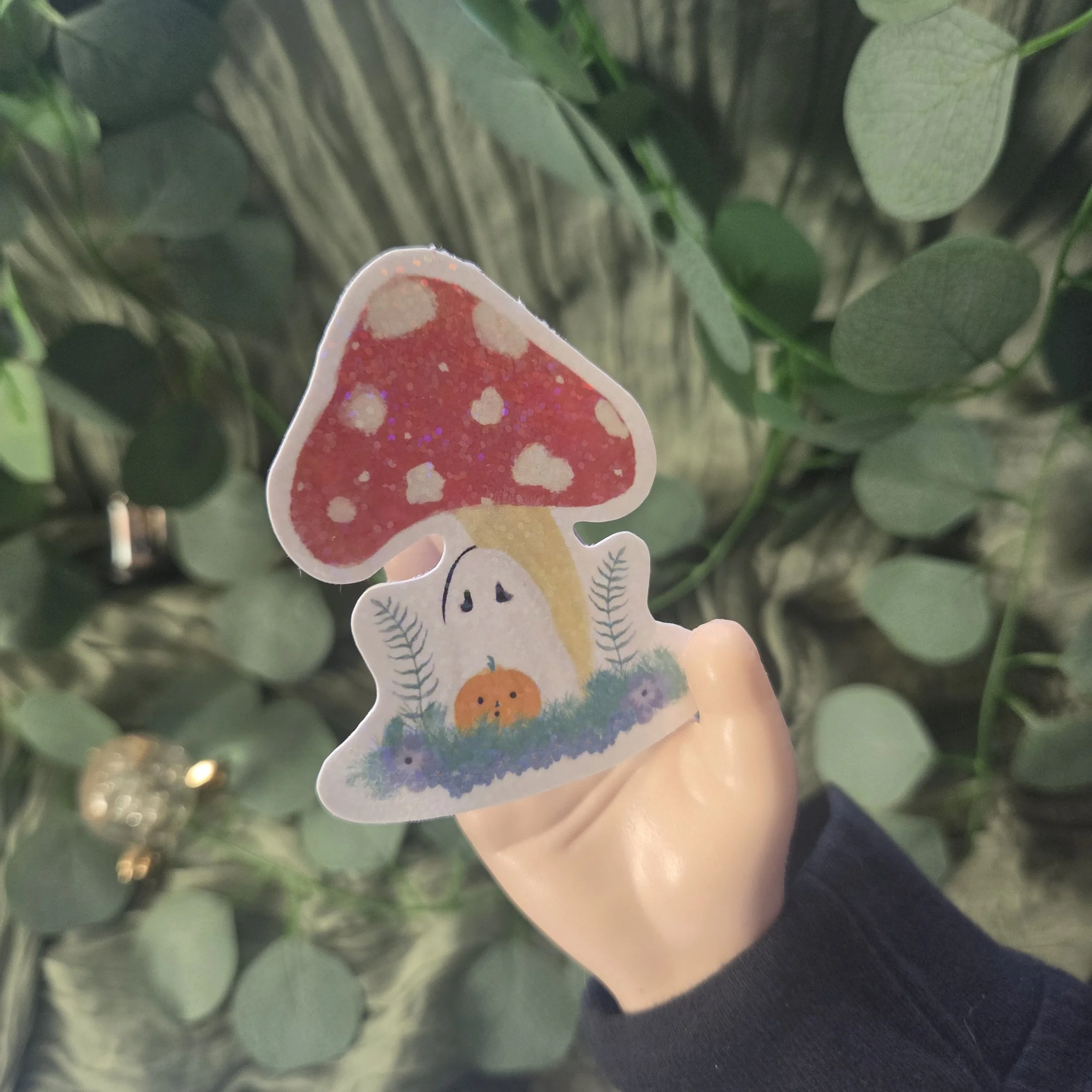 Hand holding sticker  design small ghost sitting beneath a red spotted mushroom in a whimsical autumn style. (Copy)