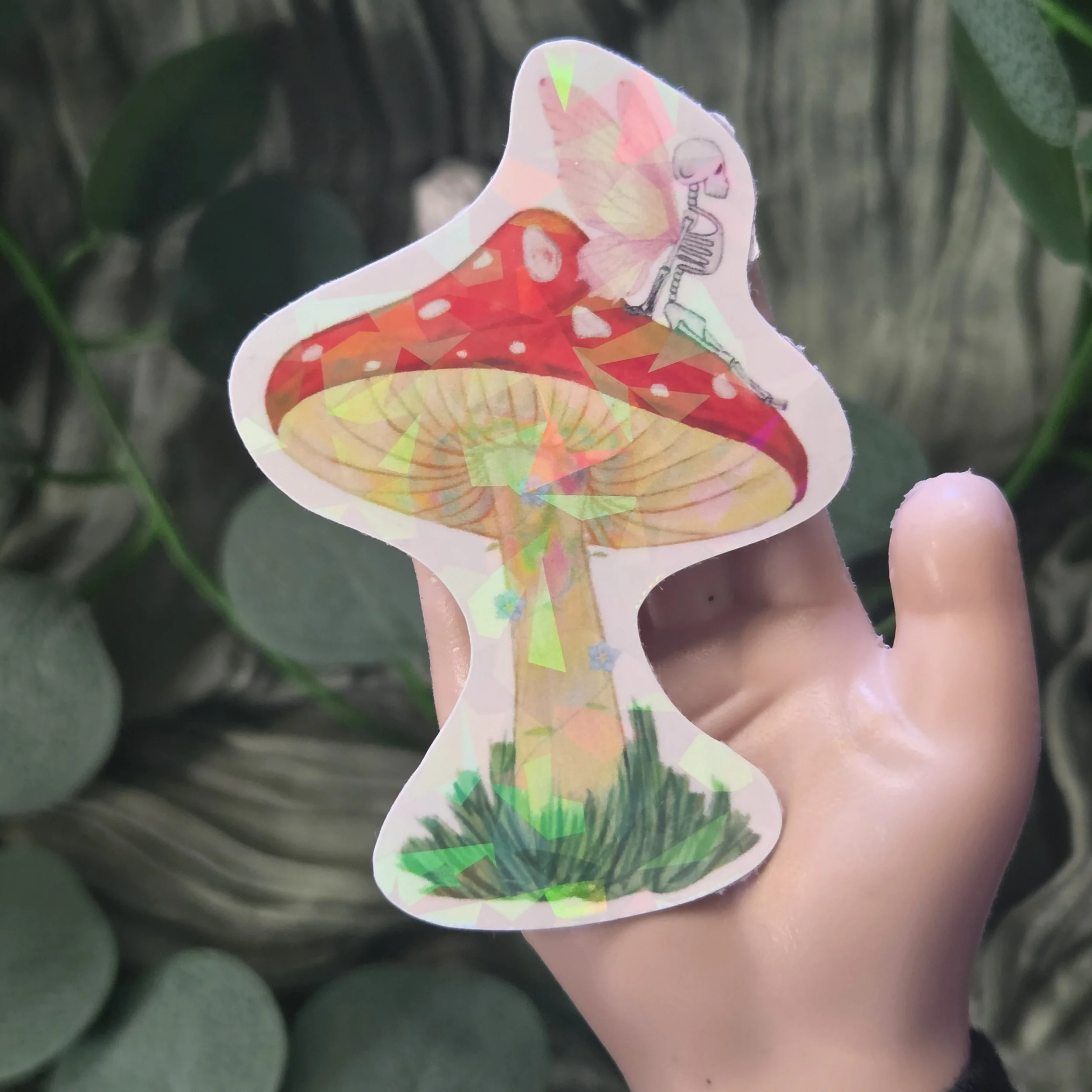 Hand holding a sticker of a skeleton fairy sitting on top of a red spotted mushroom with small wings and grass below. (Copy)