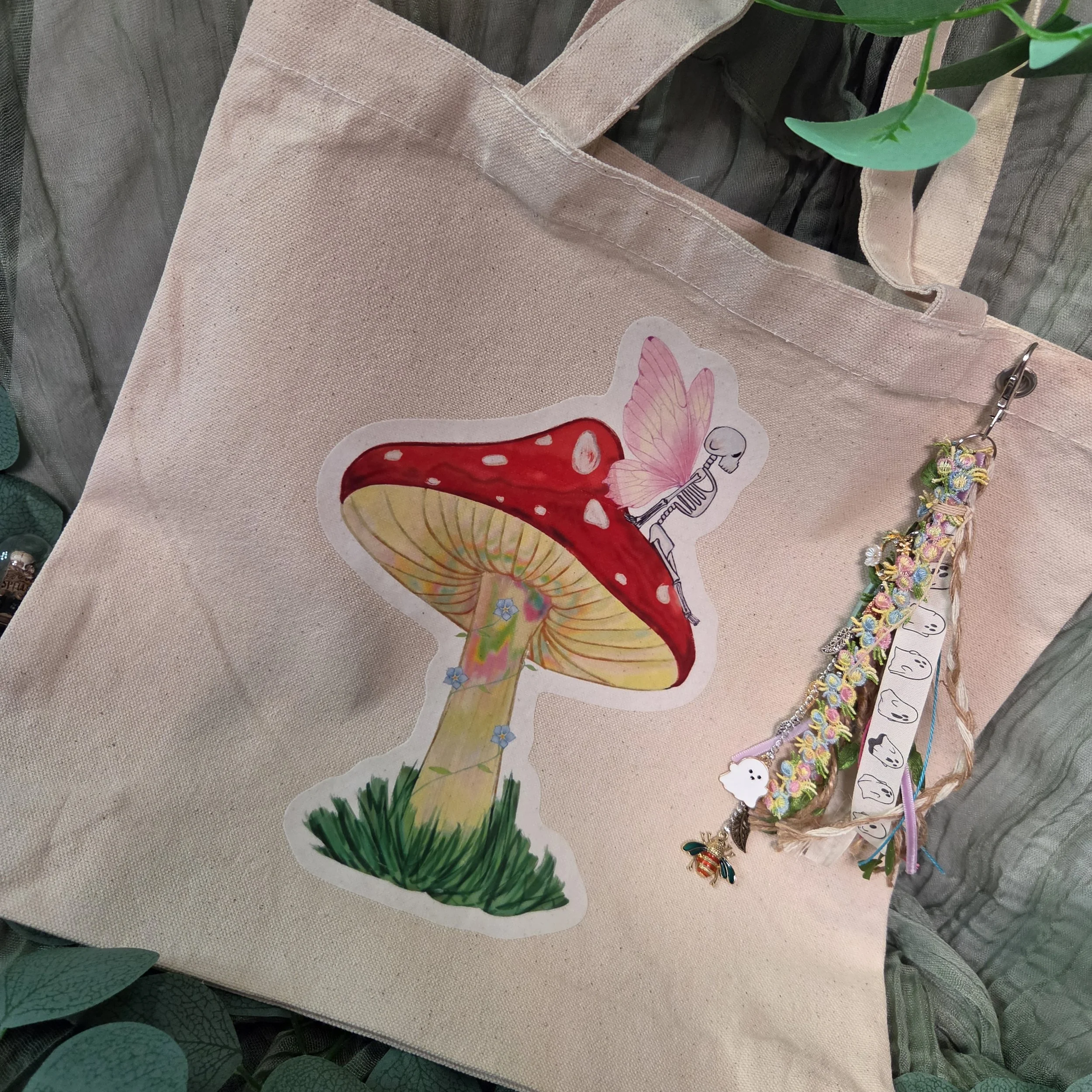 Drawing of a skeleton fairy sitting on a mushroom printed on a canvas tote bag with charm attached