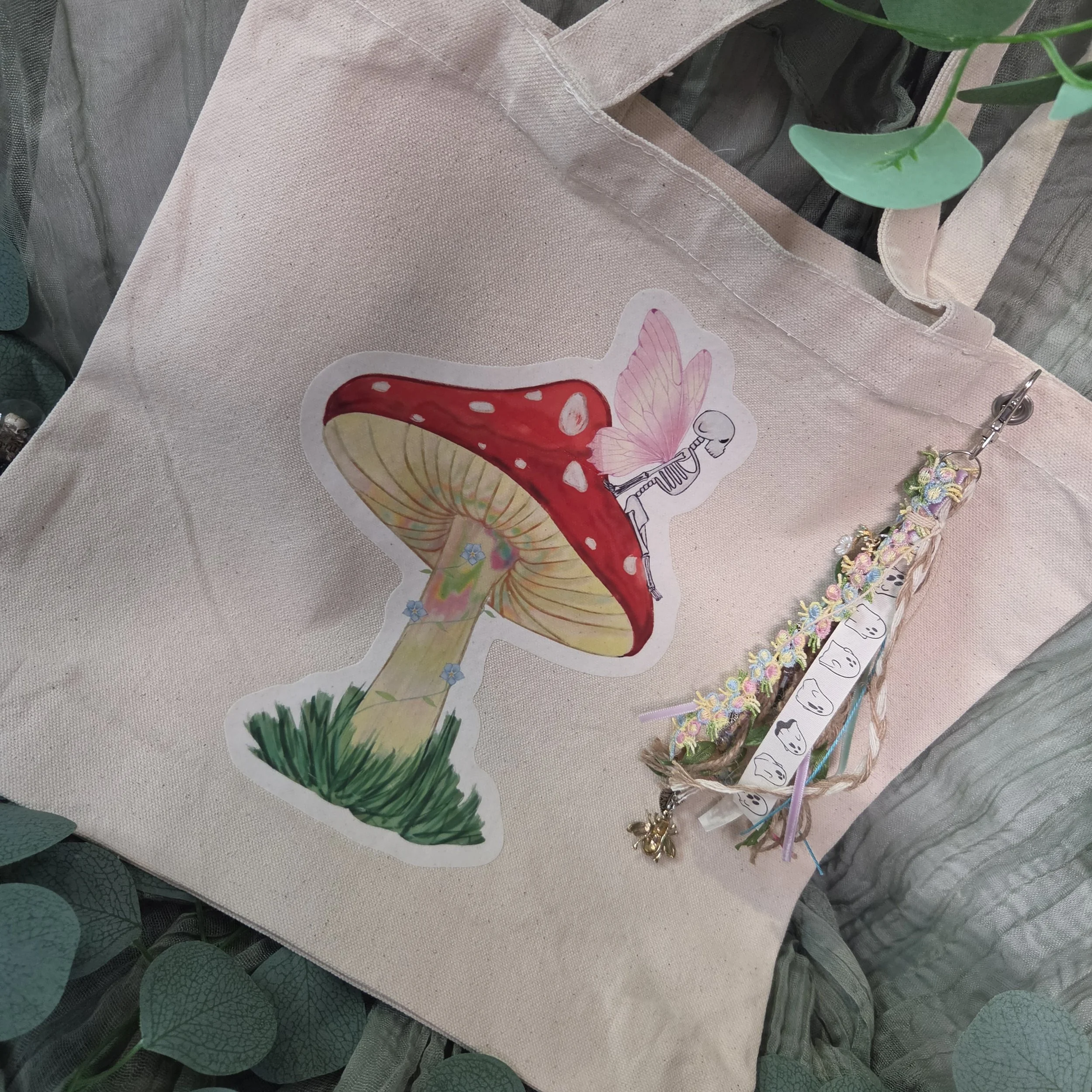 Drawing of a skeleton fairy sitting on a mushroom printed on a canvas tote bag with charm attached