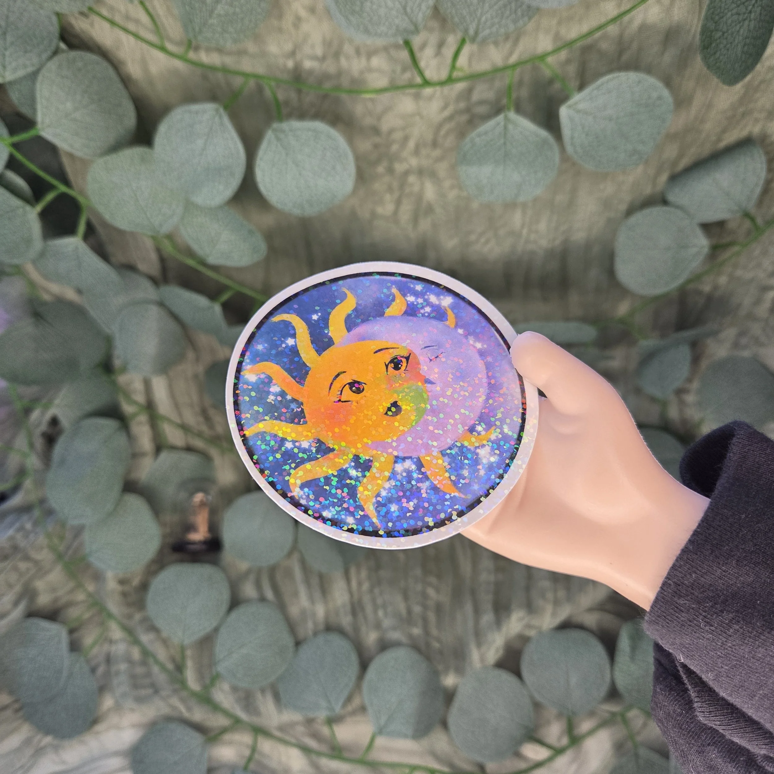 a hand holding a sticker of a drawing of a sun and moon with expressive faces intertwined against a starry night sky with a holographic laminate.