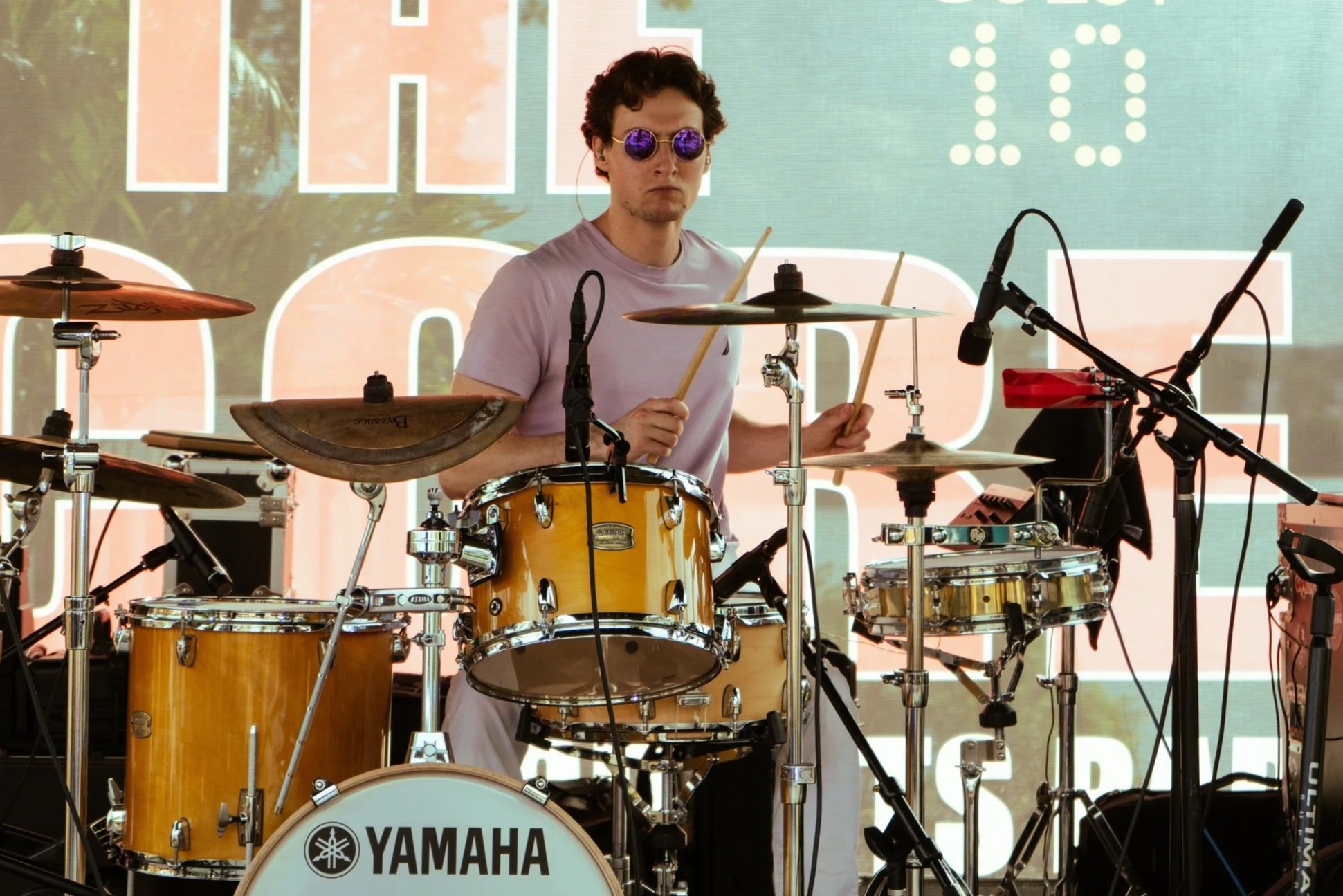 A young man wearing purple sunglasses and a white T-shirt playing a yellow Yamaha drum set on stage, with a large sign in the background reading 'THE' and 'GUEST 10'.