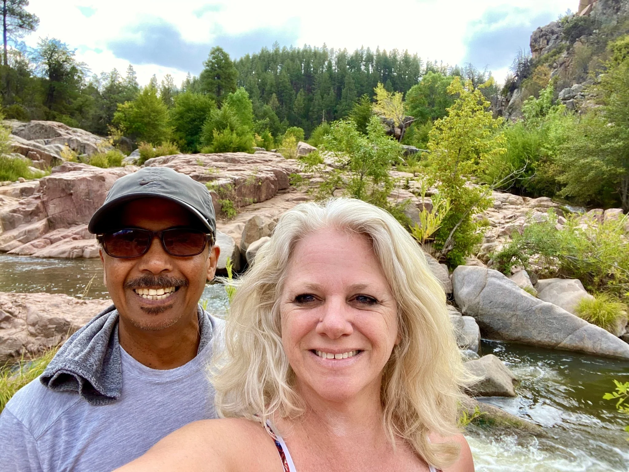 A smiling man and woman taking a selfie outdoors near a river, with rocks and green trees in the background.