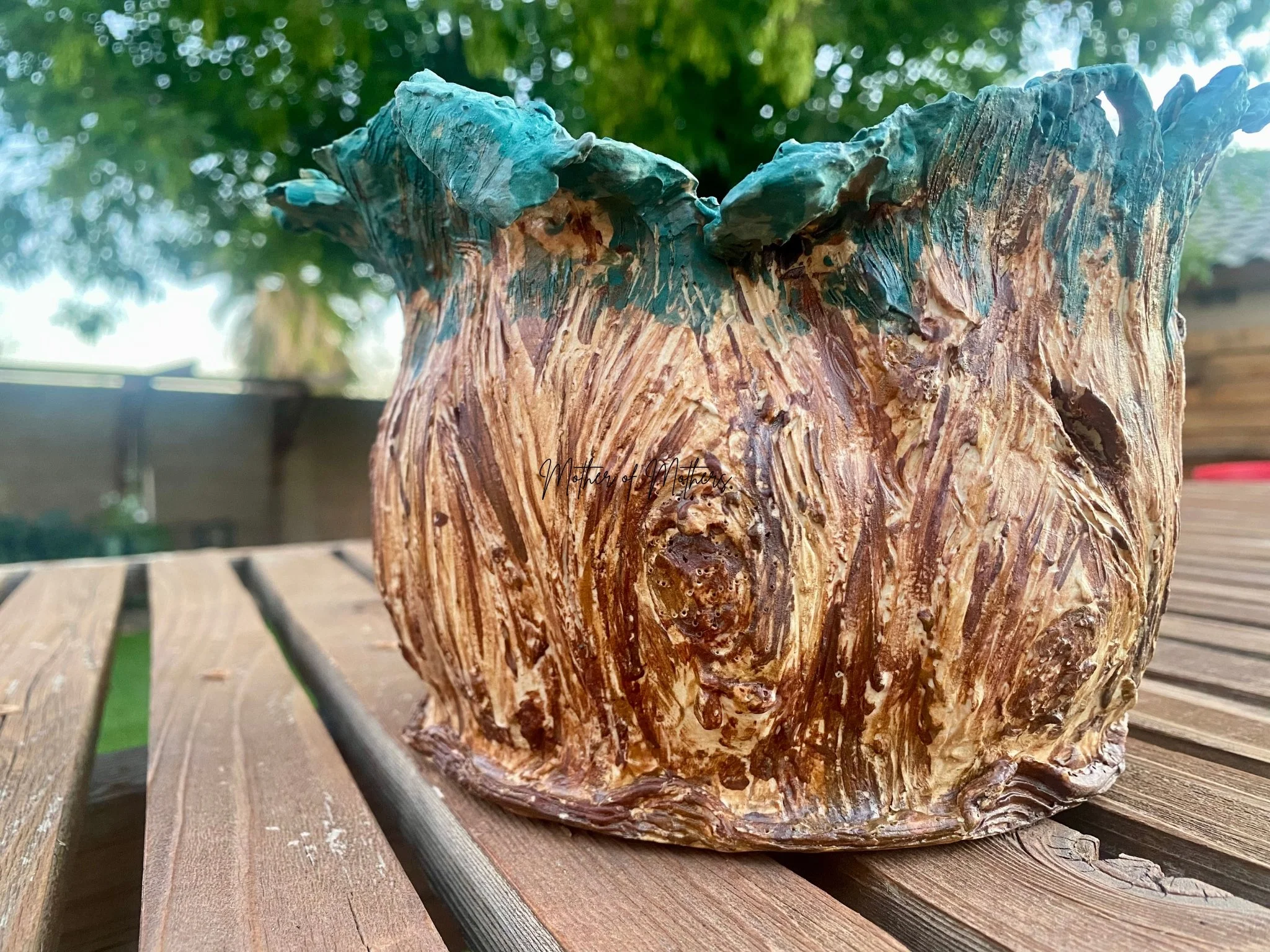 A handcrafted wooden planter with a face carved into it, featuring green painted leaves at the top, placed on a wooden surface outdoors with trees in the background.