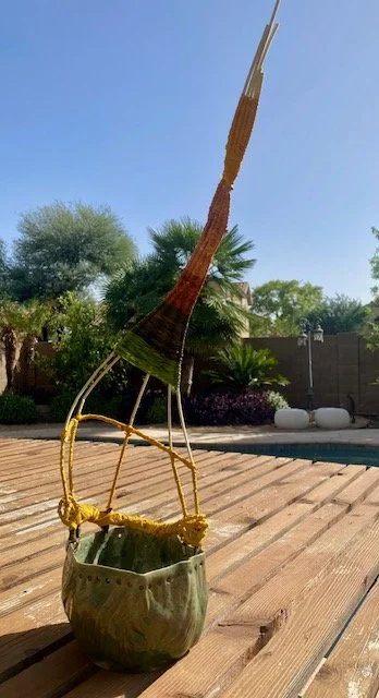 A decorative sculpture of a high-heeled shoe made from woven materials, placed on a wooden deck outdoors with trees and a pool in the background.