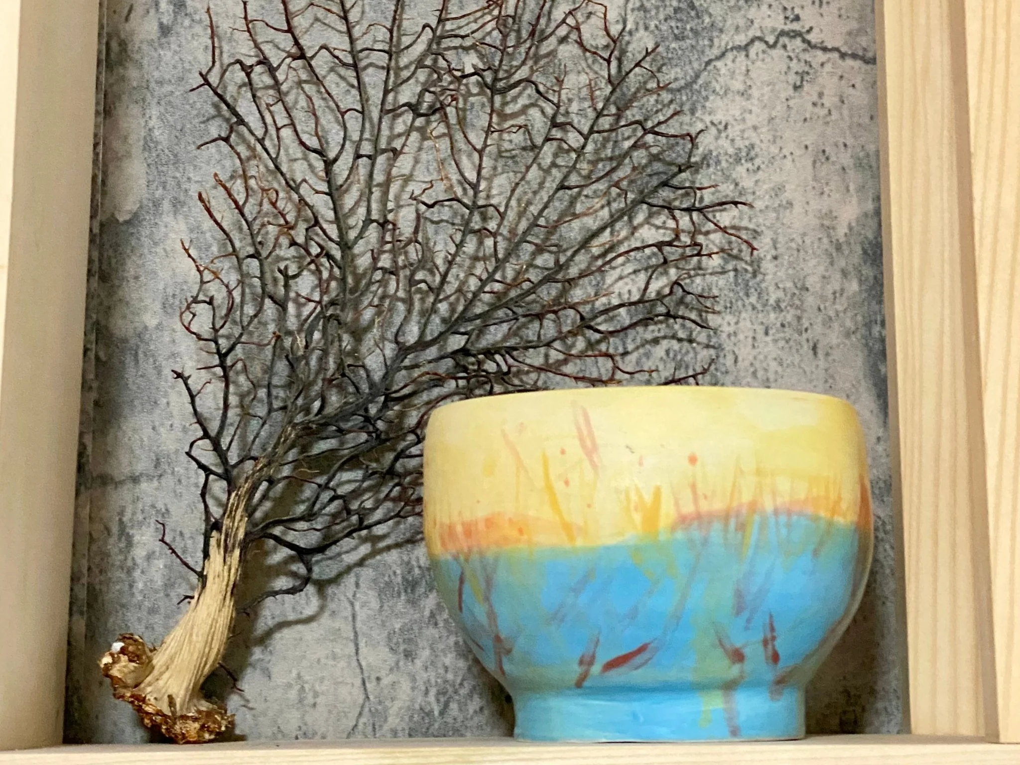A decorative arrangement featuring a bare, leafless tree branch and a colorful ceramic bowl on a wooden shelf with a textured grey background.