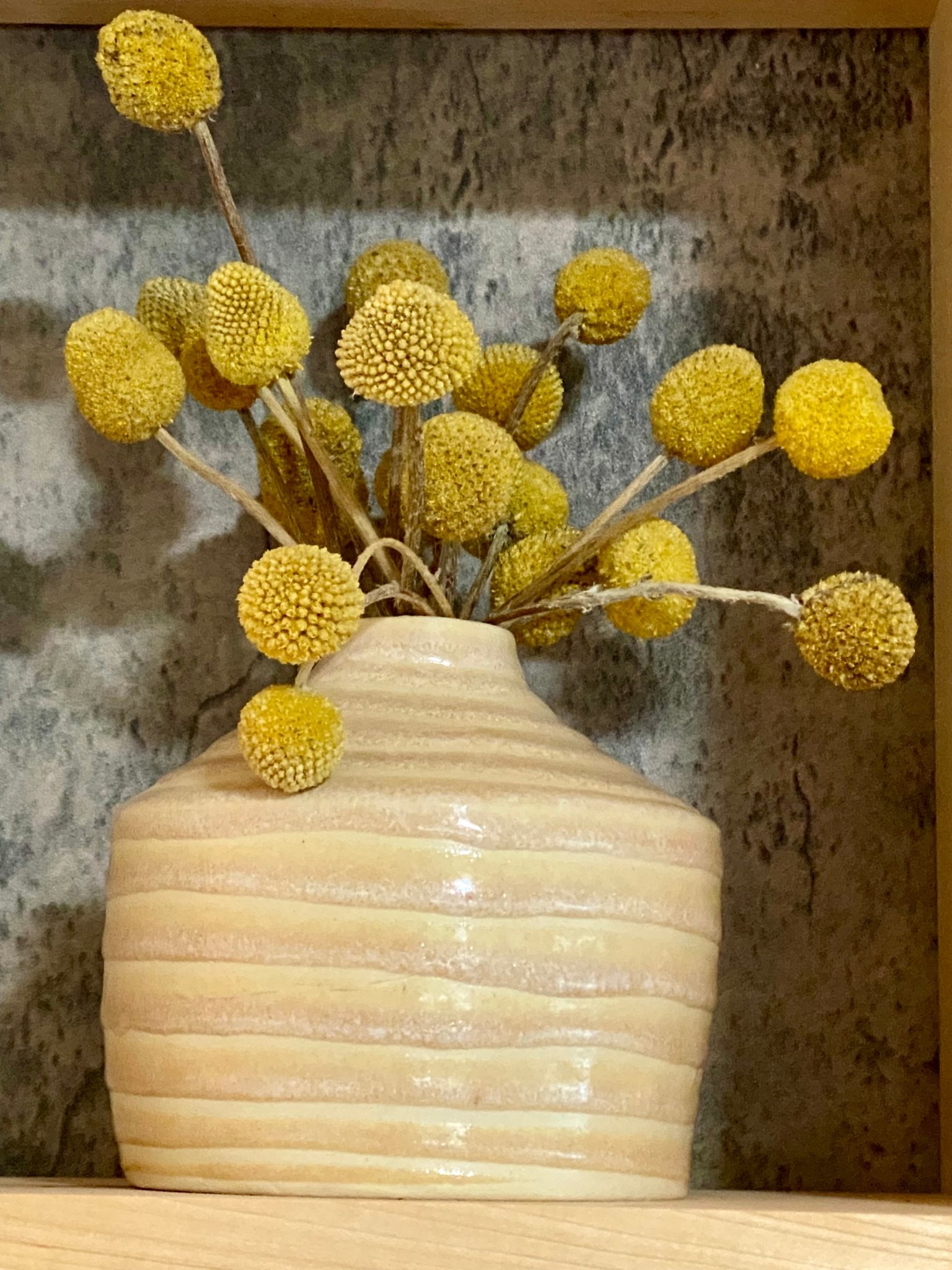 Yellow dried flower stems in a beige striped ceramic vase against a textured wall.