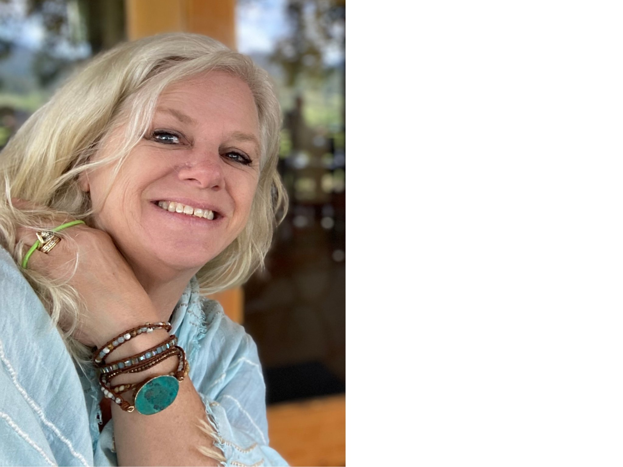 Smiling woman with blonde hair, wearing layered bracelets and a ring, resting her hand on her neck, indoors with a blurred background.