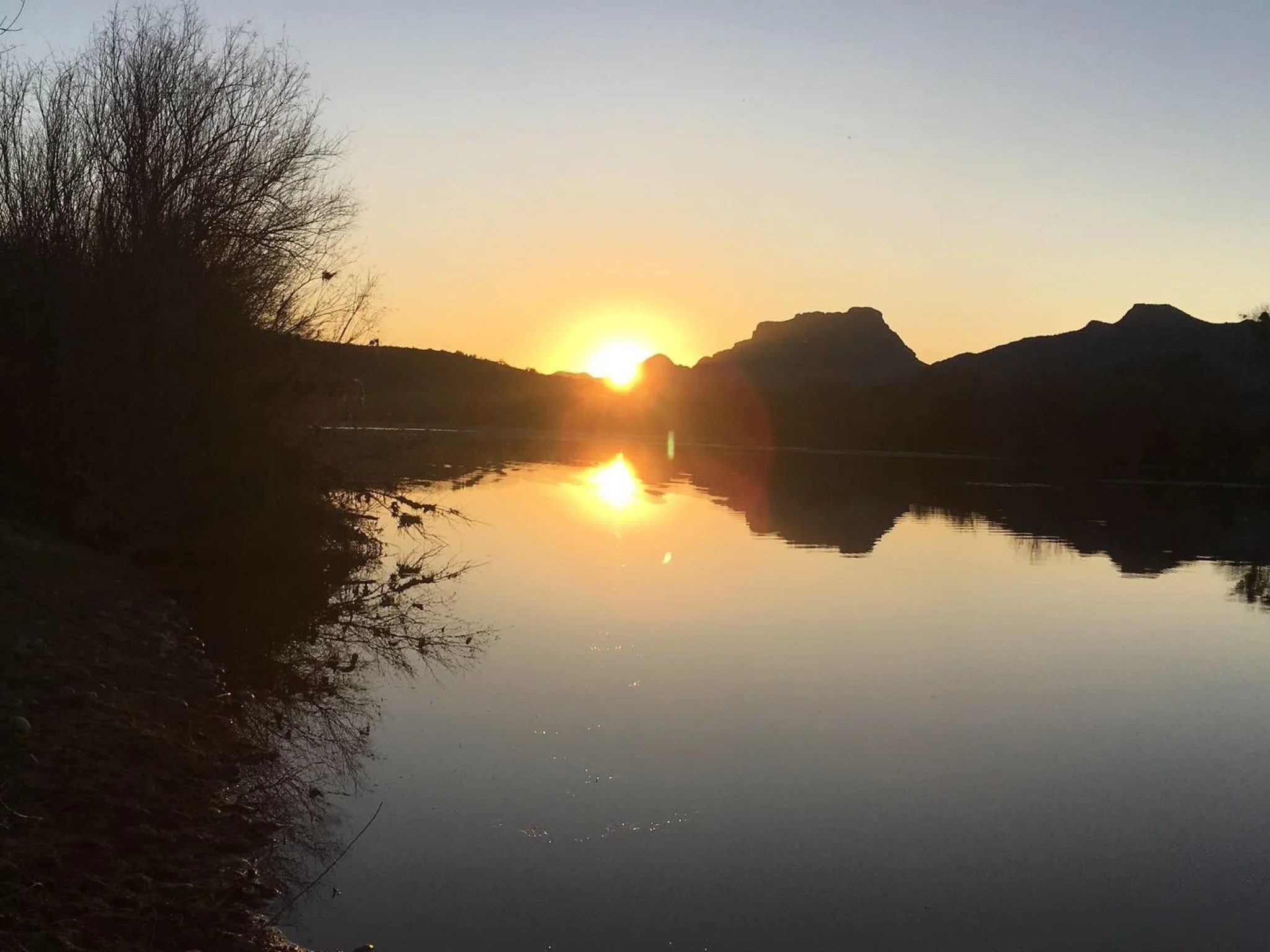 Sun setting over a river with mountains in the background and a bush on the left side.