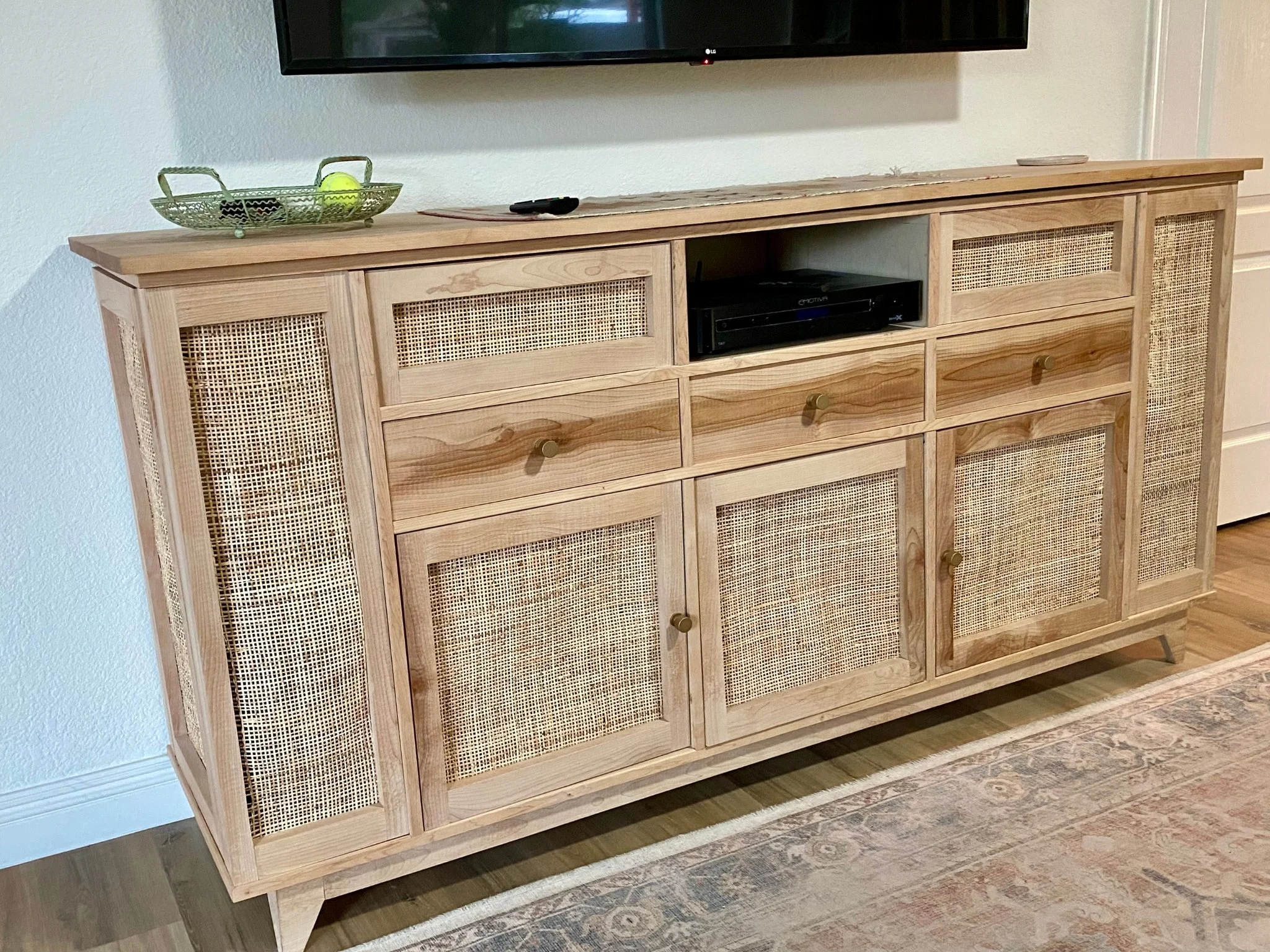 Wooden TV console with rattan cabinet doors, a central open shelf with electronic devices, and a decorative tray with a tennis ball on top, situated on a hardwood floor next to a patterned rug.