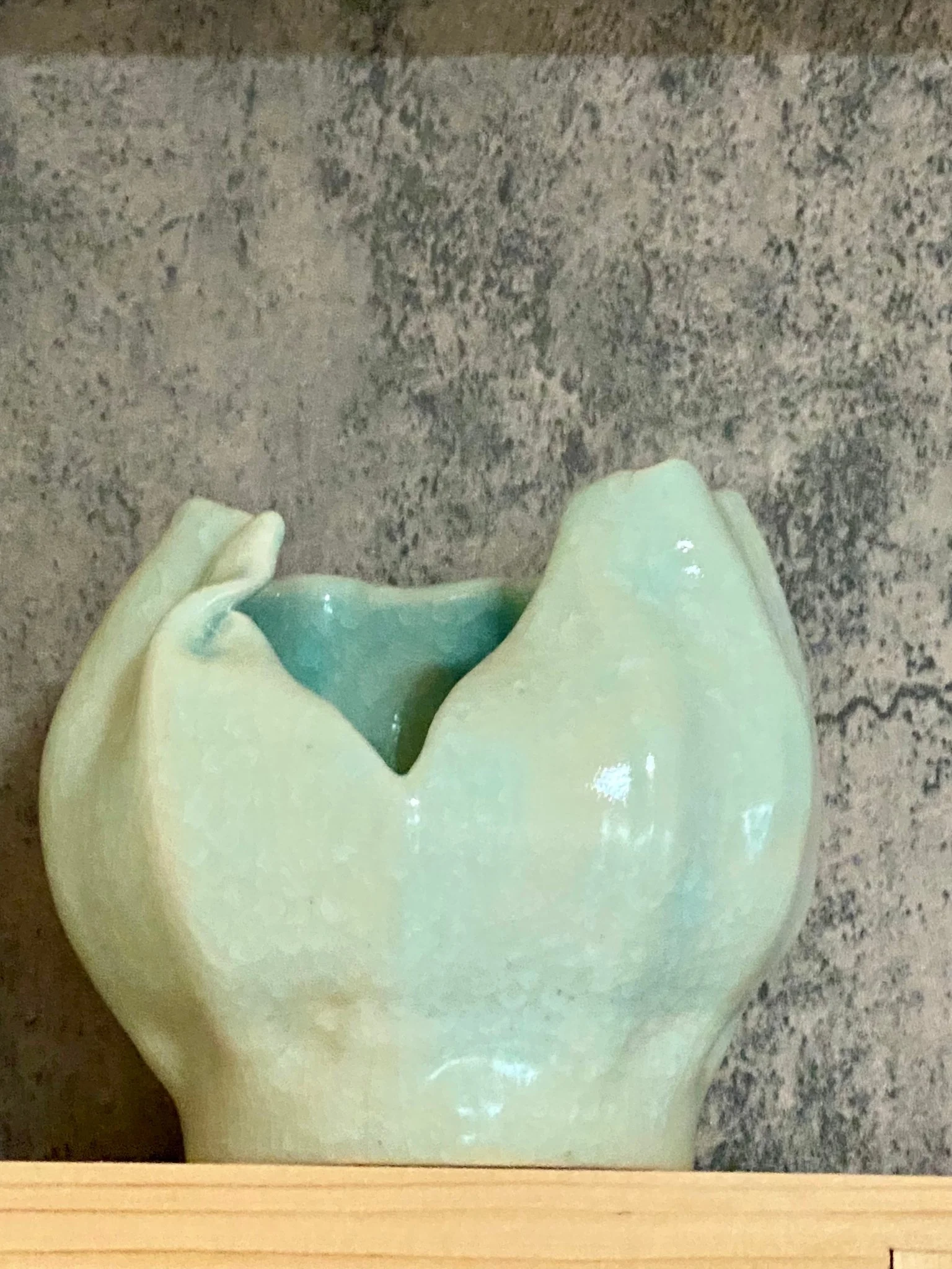 A broken jade vase with a light green exterior and a blue interior, sitting on a wooden surface against a gray wall.