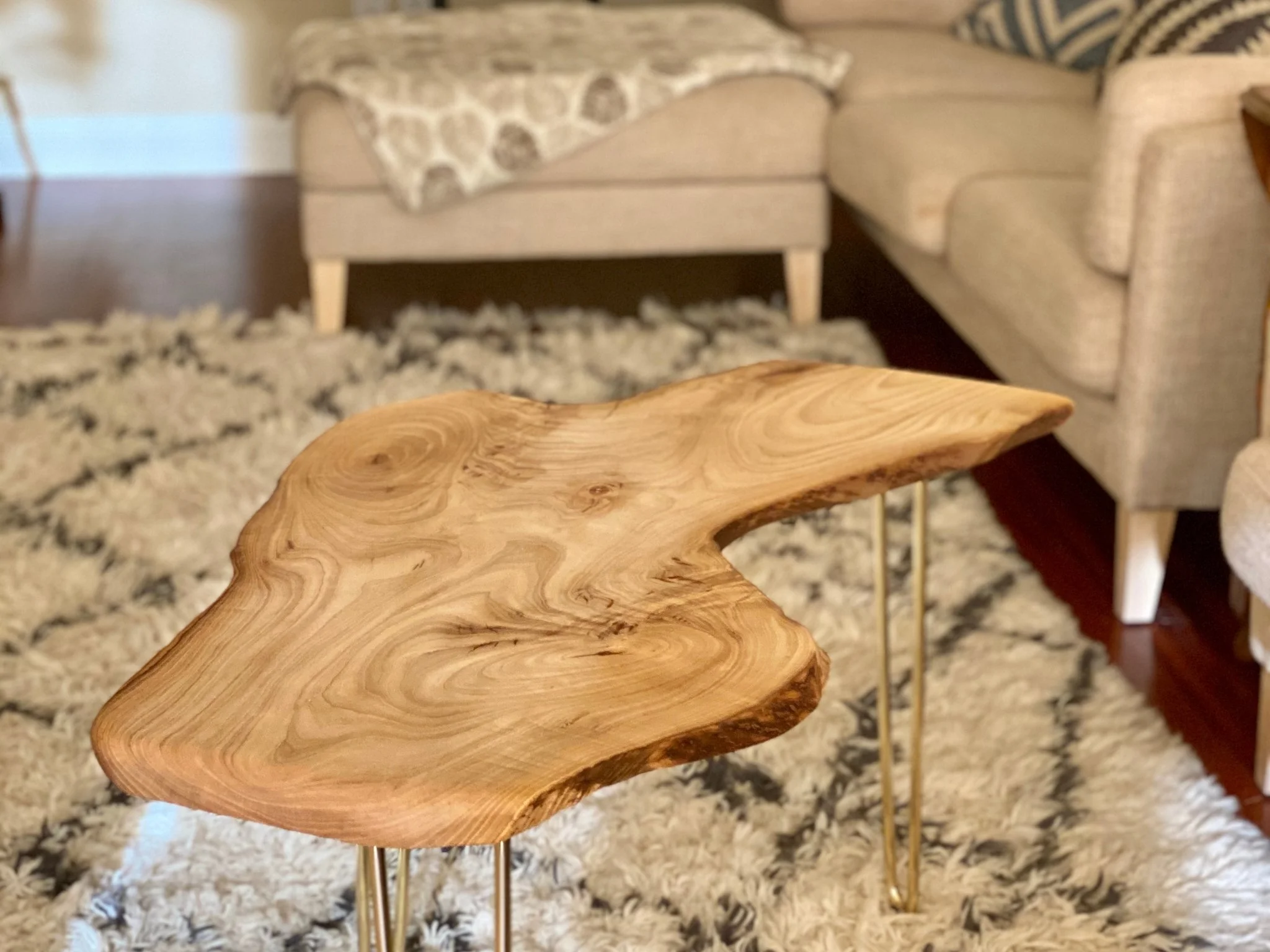 Close-up of a wooden side table with a natural, irregular shape and visible wood grain in a living room setting with a patterned rug, beige sofa, and other furniture.